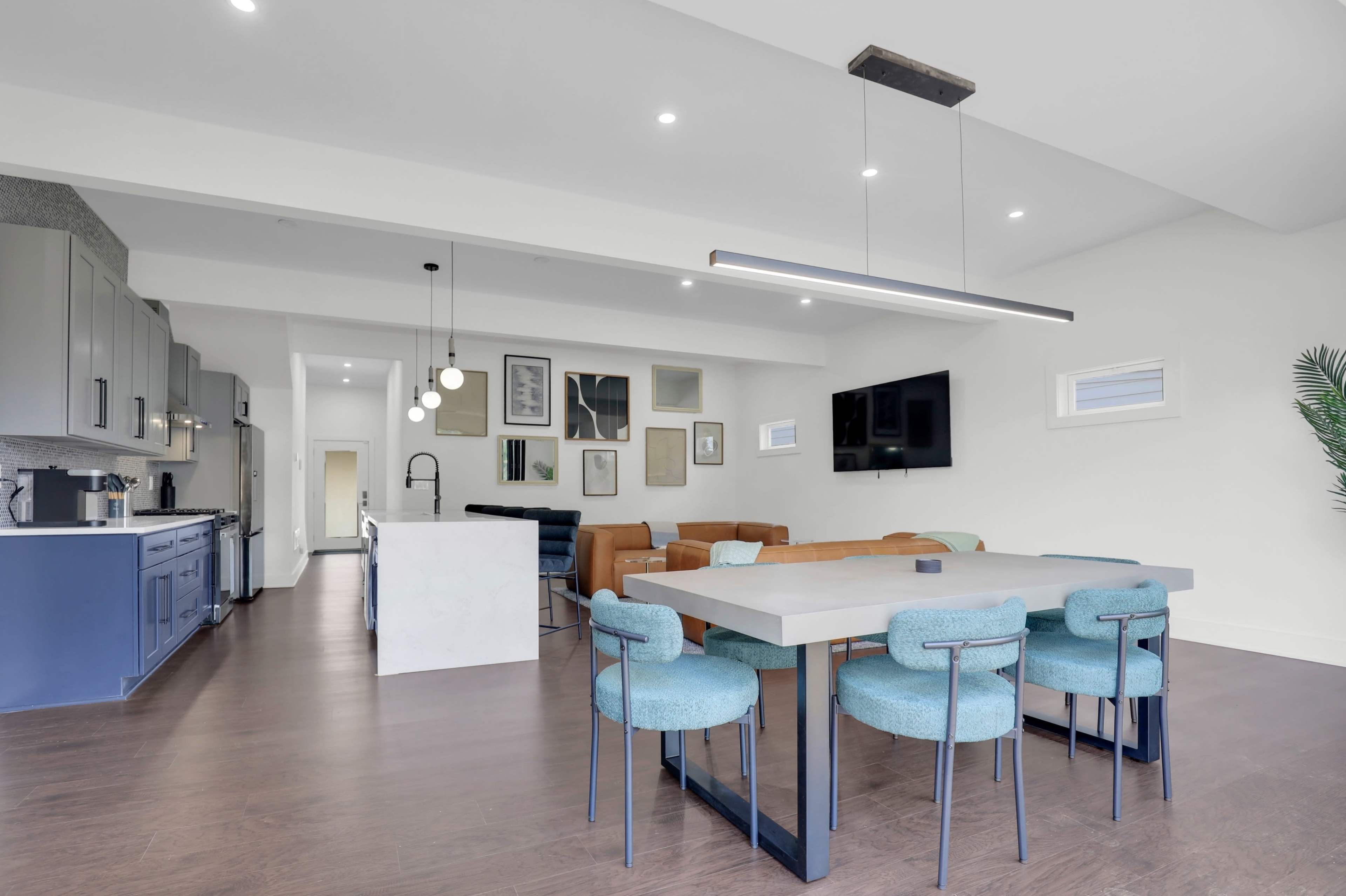 The image shows a modern open-concept living space featuring a dining table with chairs, a sectional sofa, a television, and various framed artworks on the walls.