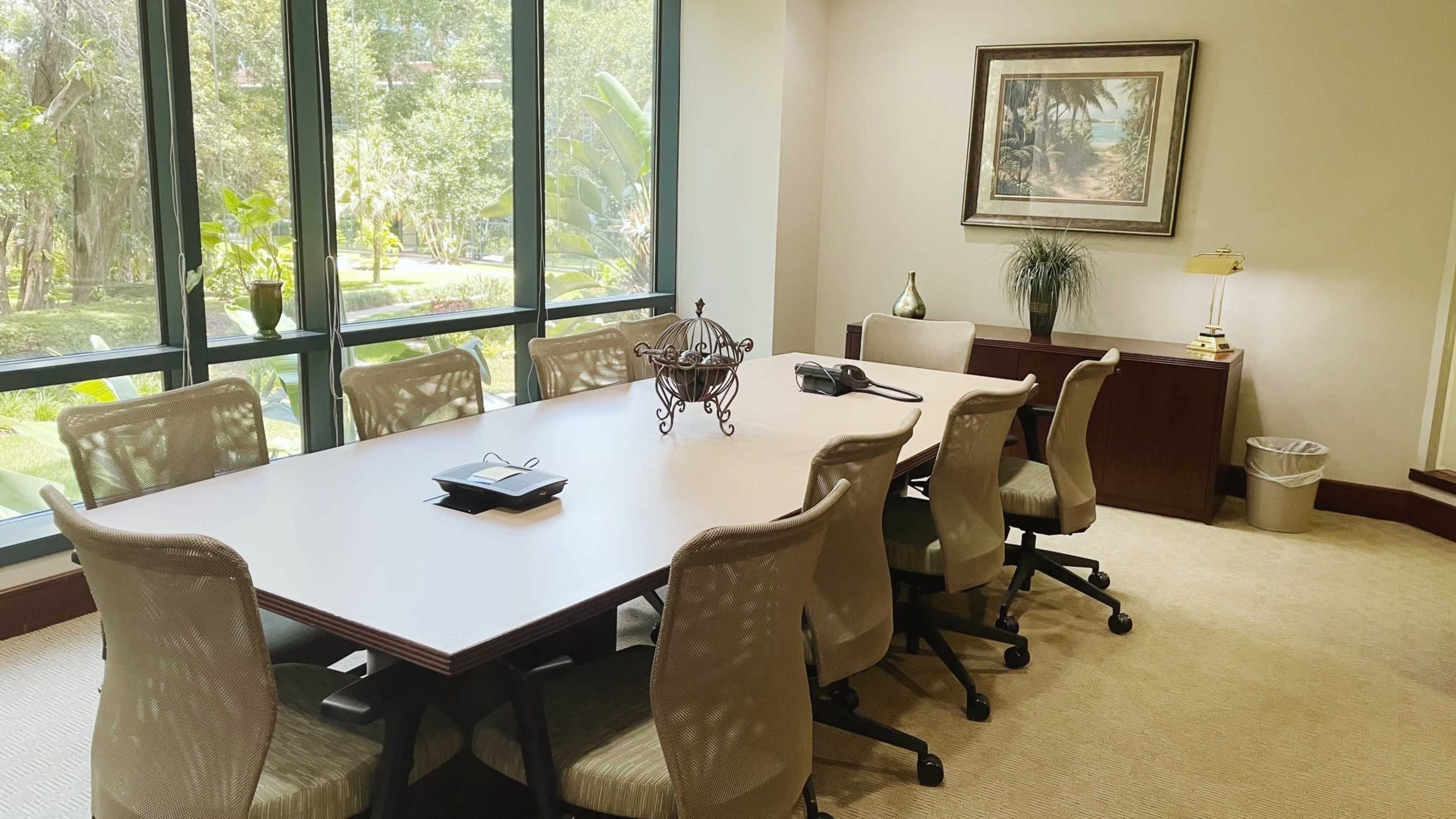 A conference room features a large table surrounded by several chairs, with a phone and decorative items on the table, and large windows overlooking greenery.