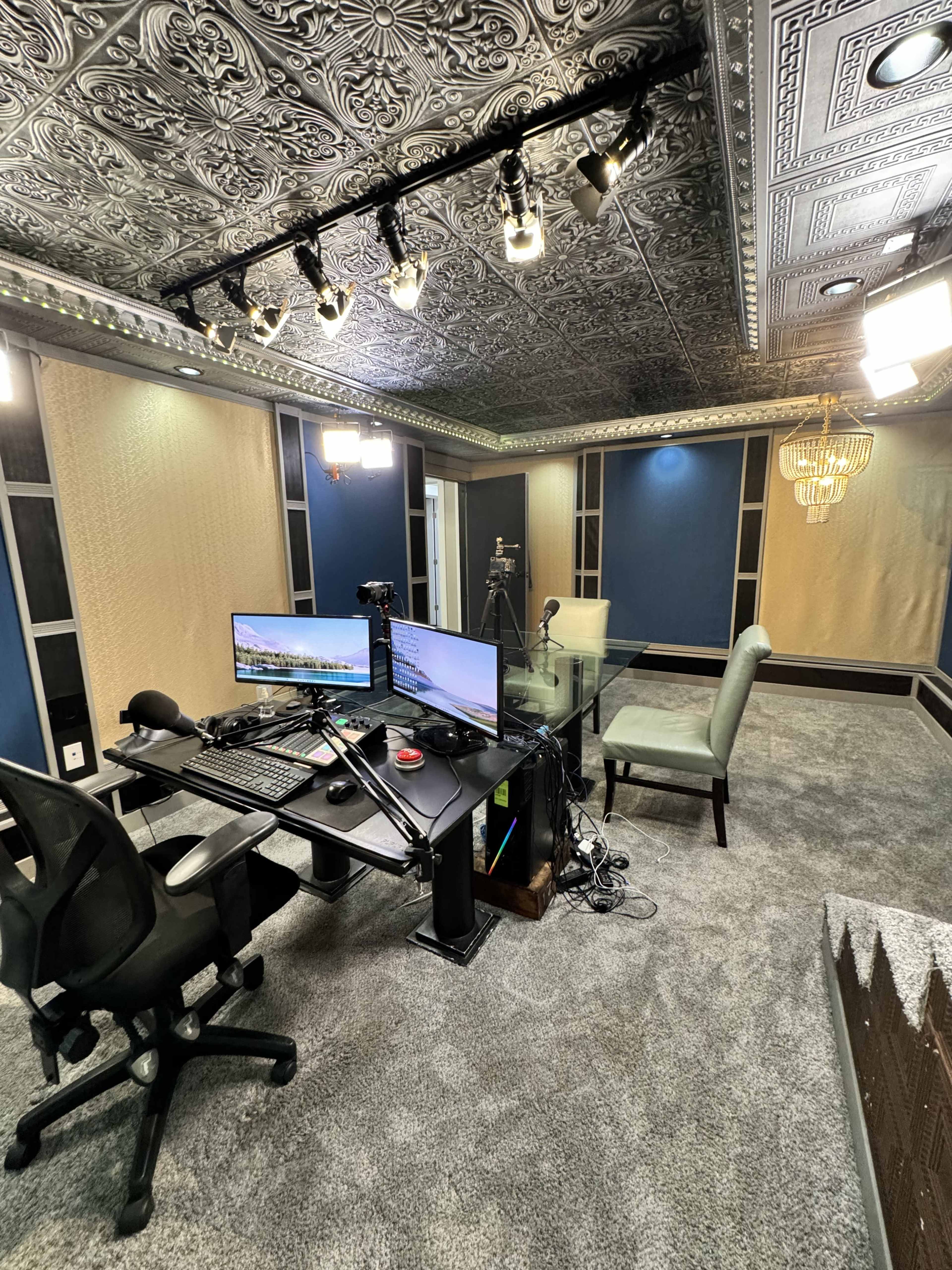 The image shows a modern recording studio with two computer monitors, a microphone, and a glass table surrounded by plush carpeting and ornate ceiling details.