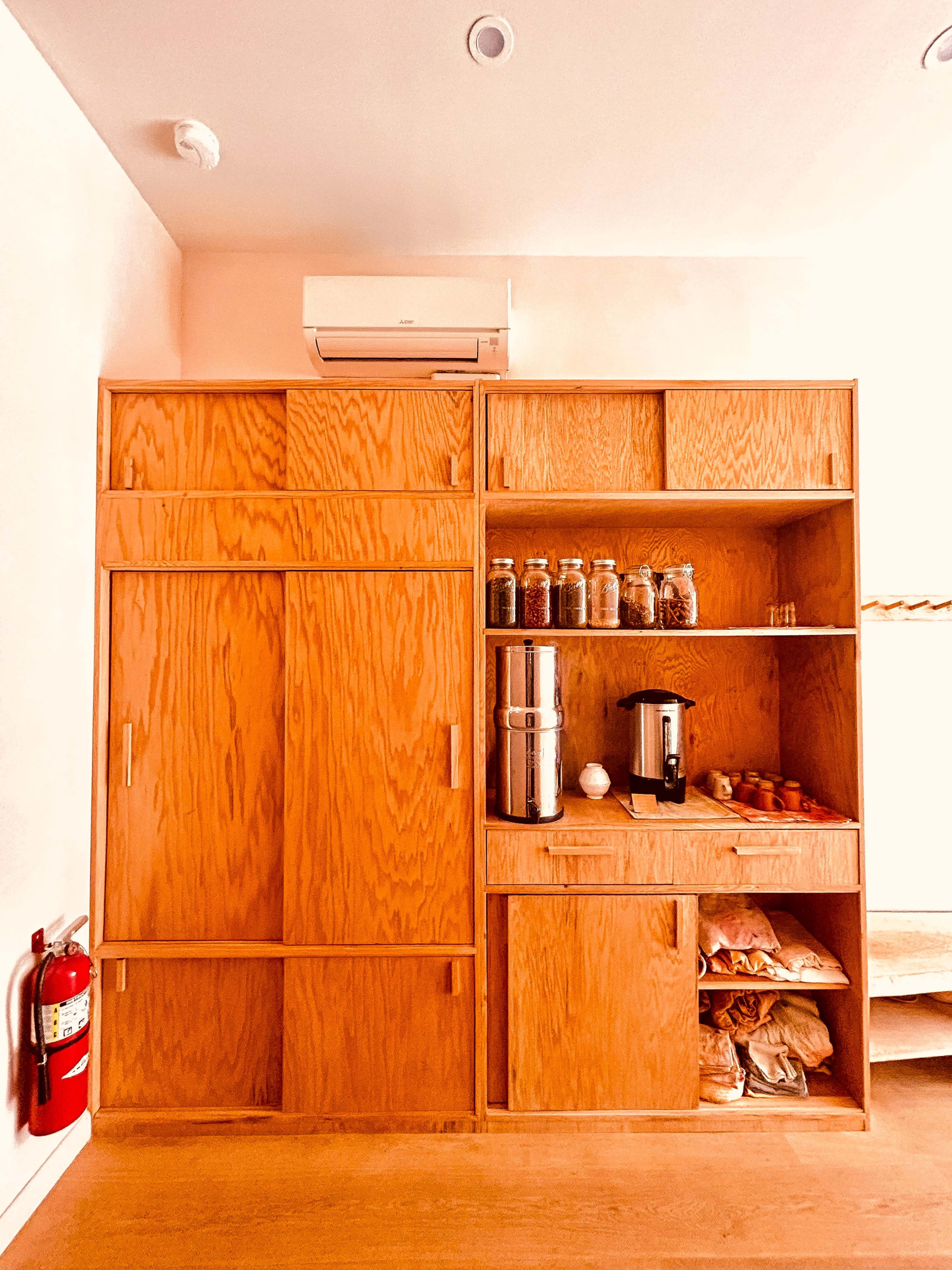 The image shows a wooden storage unit with shelves containing jars, a coffee maker, and bags of items, alongside a fire extinguisher mounted on the wall.