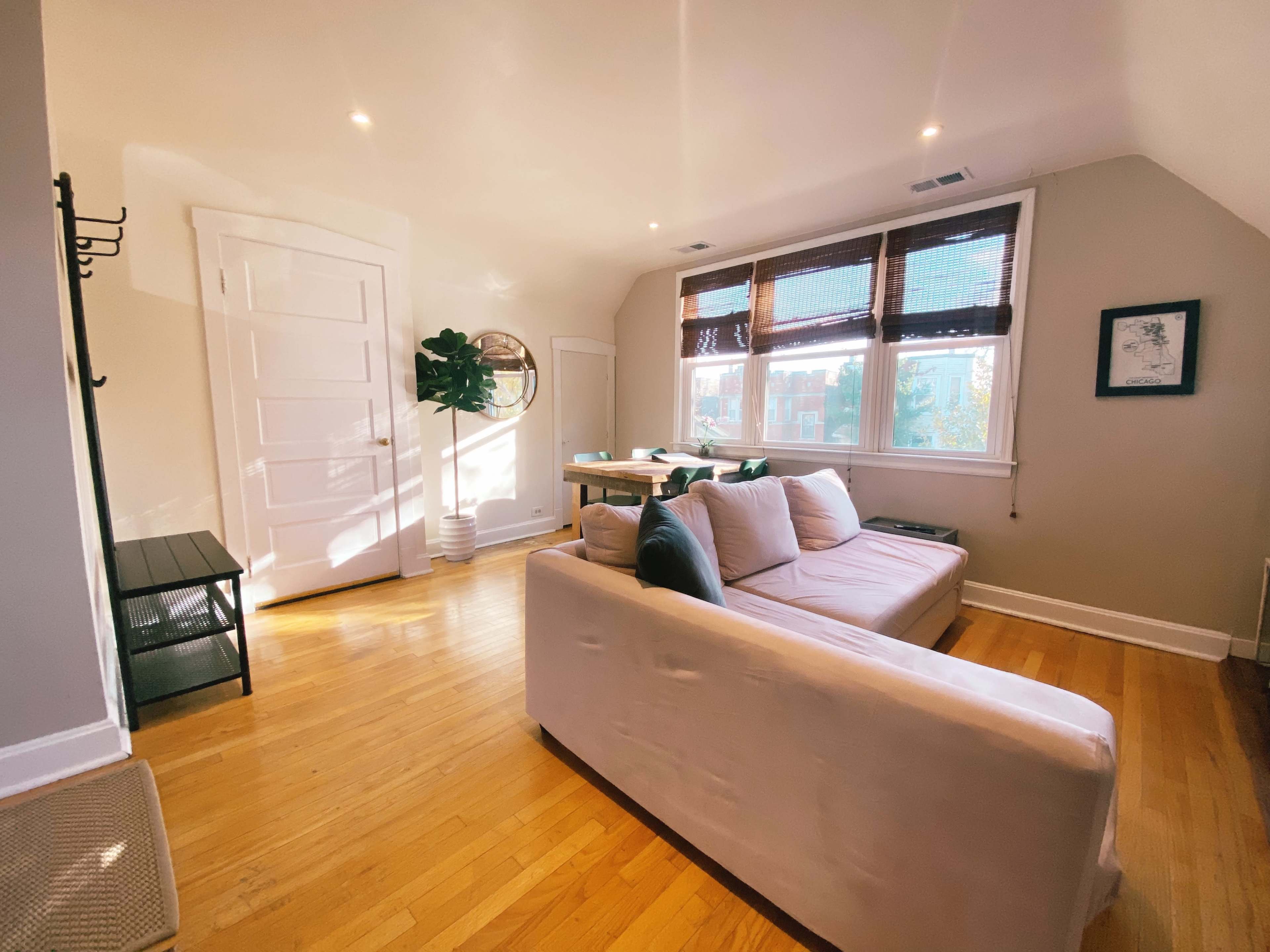 The image shows a cozy living room with a white couch, wooden flooring, and large windows covered by bamboo shades.