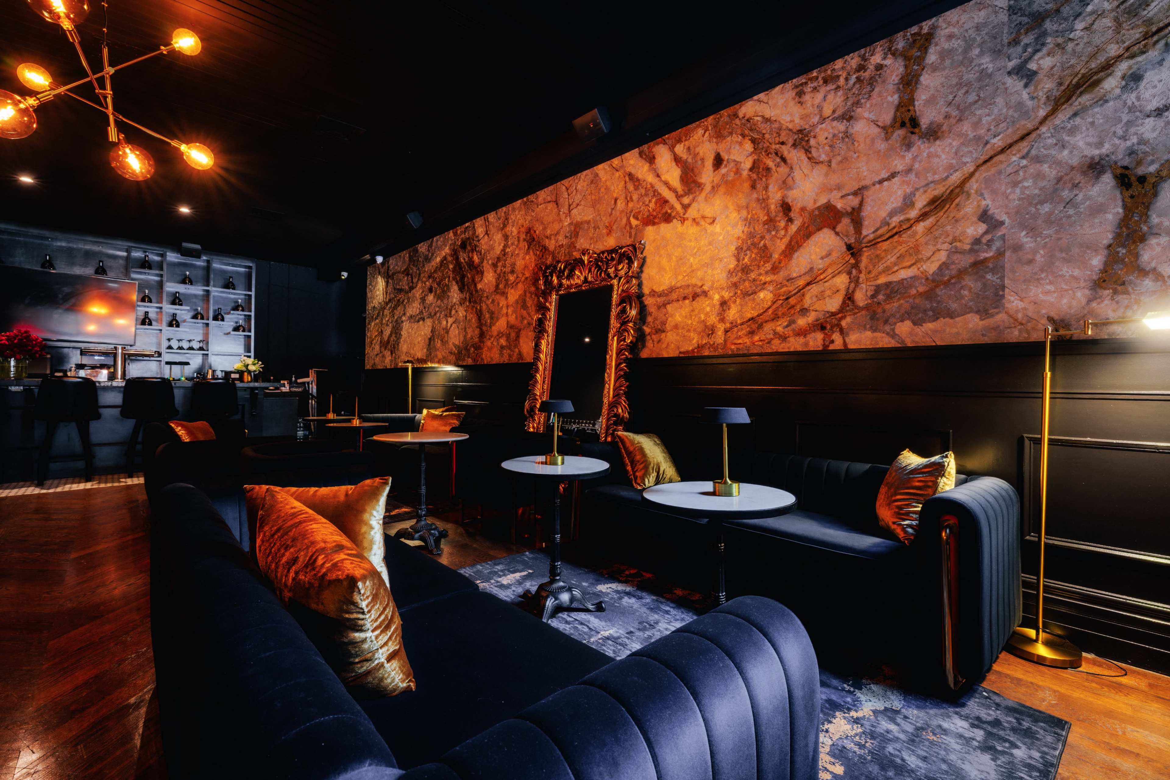 The image features a stylish lounge area with dark walls, elegant furniture, and a large mirror framed in gold.