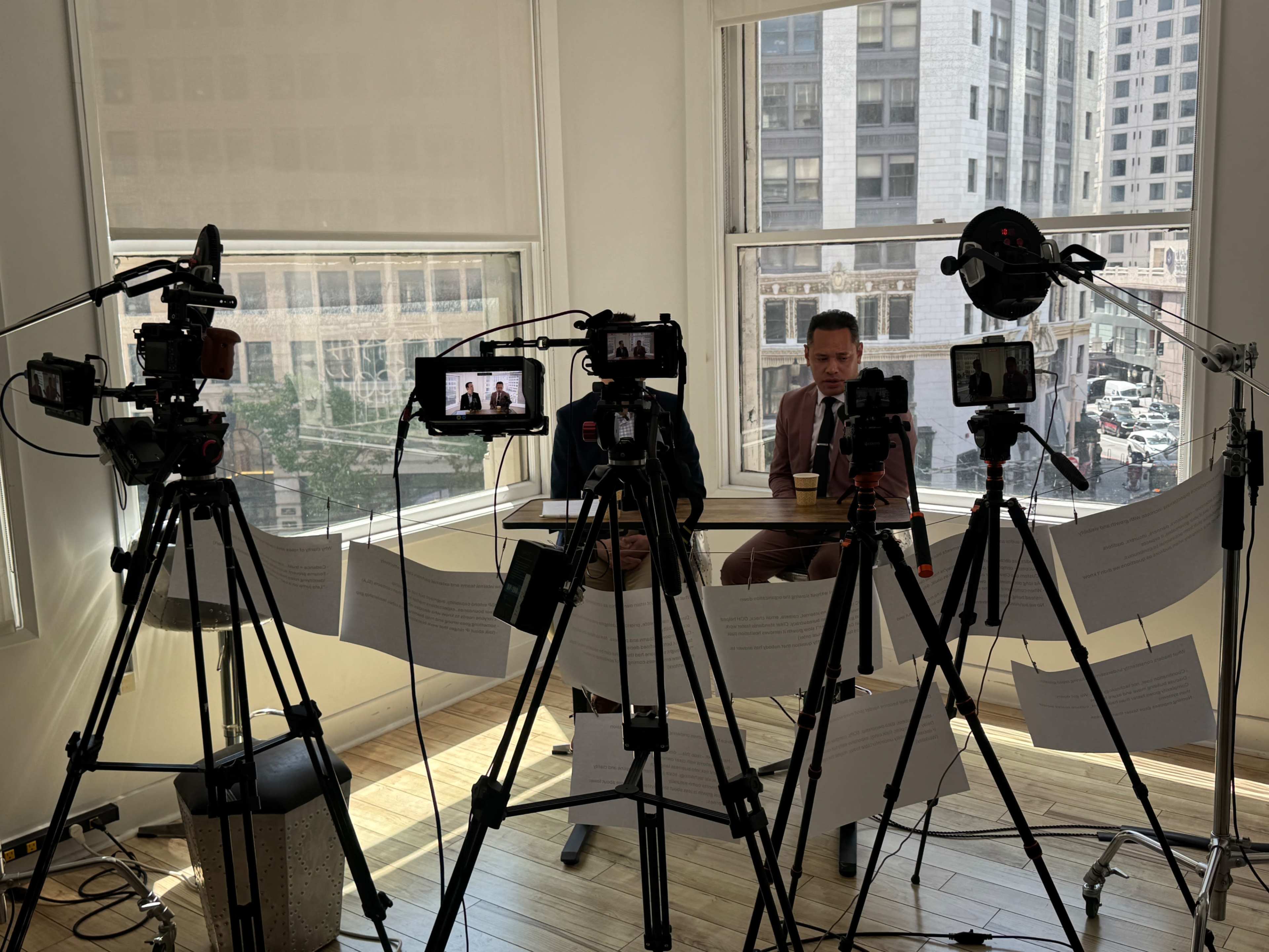 A man sits at a table in a well-lit room, surrounded by multiple cameras and microphones, with urban buildings visible through the large windows behind him.