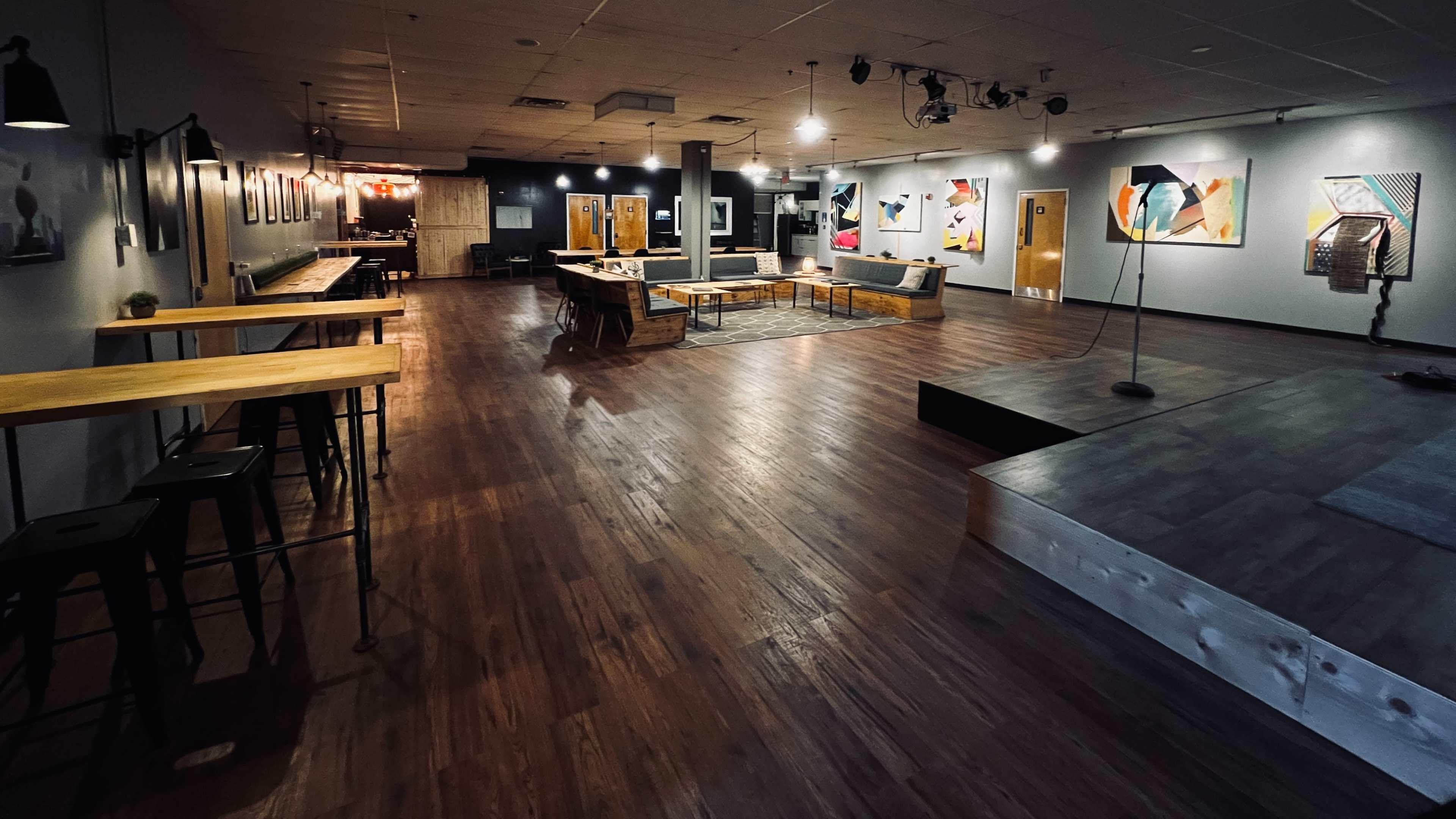 A spacious interior venue with wooden floors, various artworks on the walls, a stage area, and seating arrangements including tables and chairs.