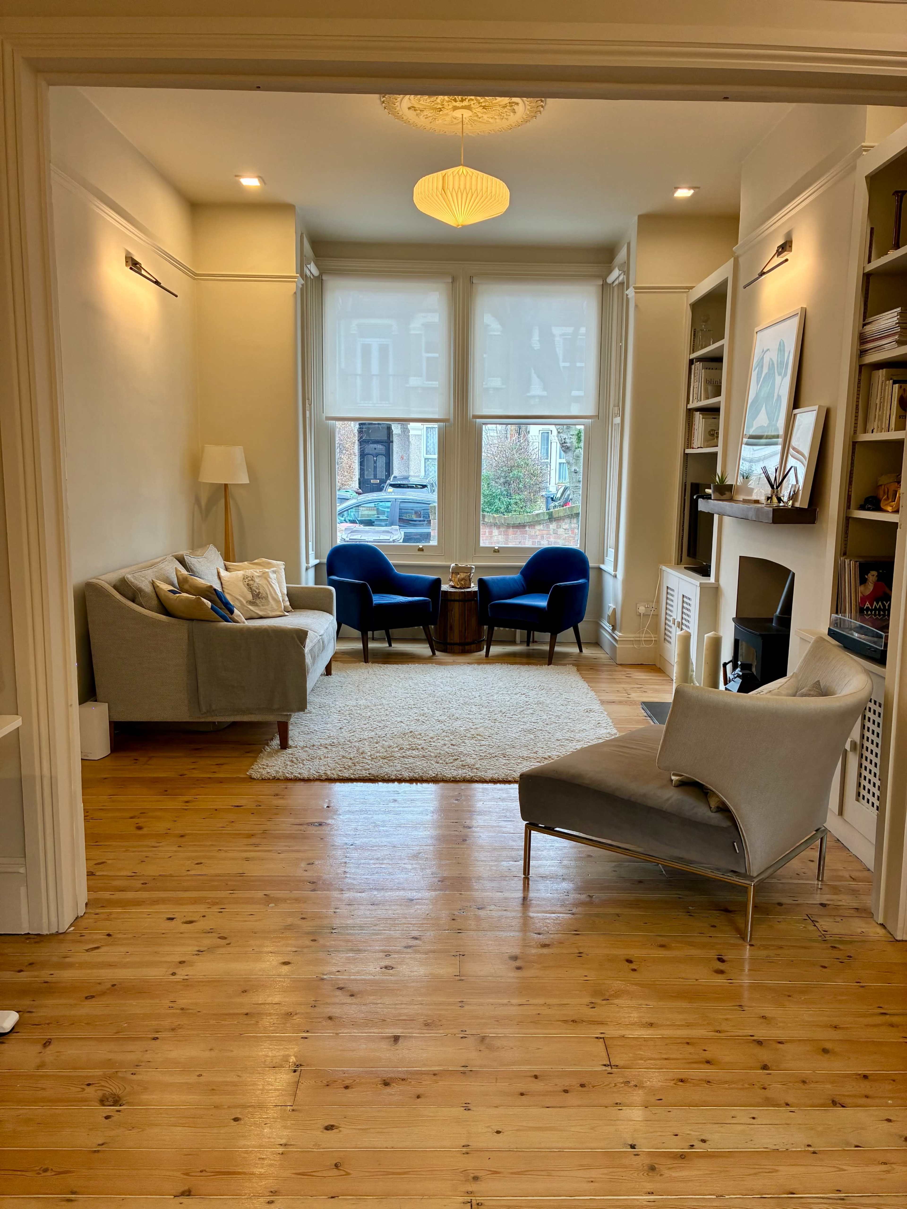 The image shows a cozy living room featuring two blue chairs, a small wooden coffee table, and a light-colored rug, with large windows allowing natural light to enter.
