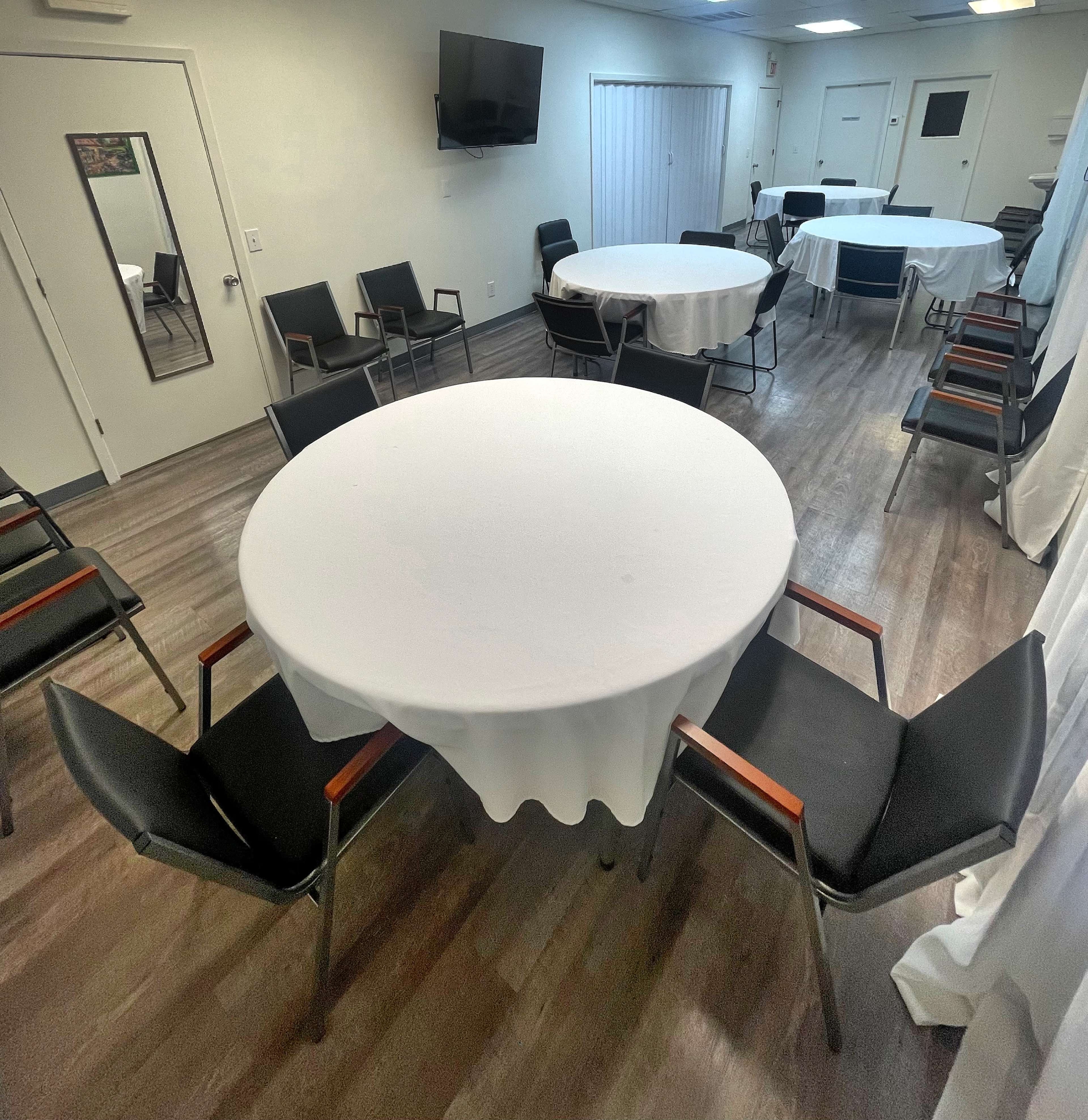 The image shows a meeting room with circular tables covered in white tablecloths arranged around black chairs, and a wall-mounted television in the background.