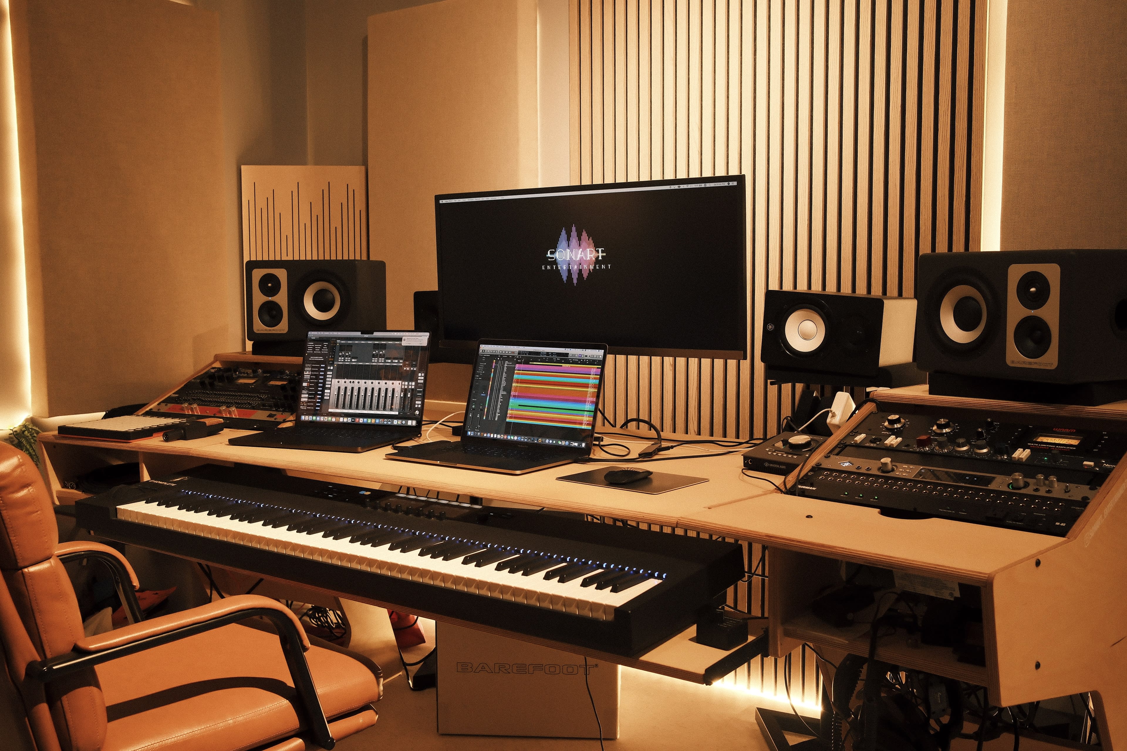 The image shows a music production studio with a keyboard, two laptops, audio monitors, and various music equipment arranged on a wooden desk.