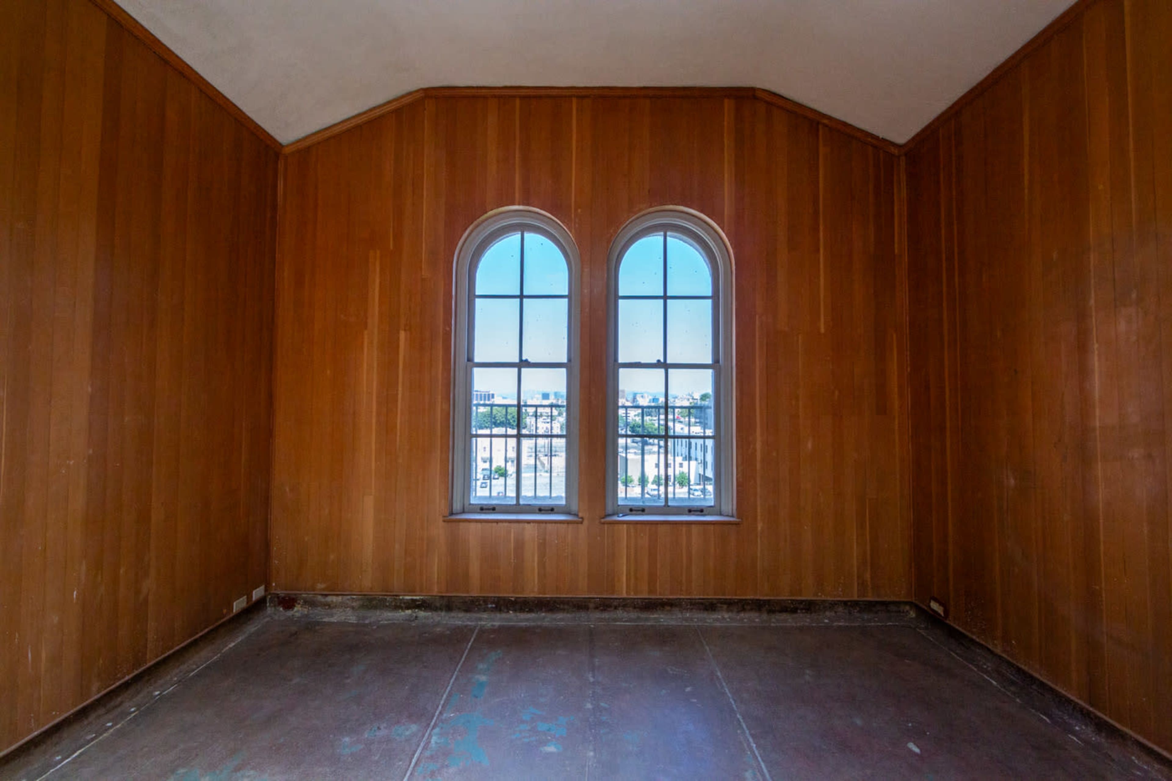 The image shows an empty room with wooden paneling and two arched windows that overlook a cityscape.