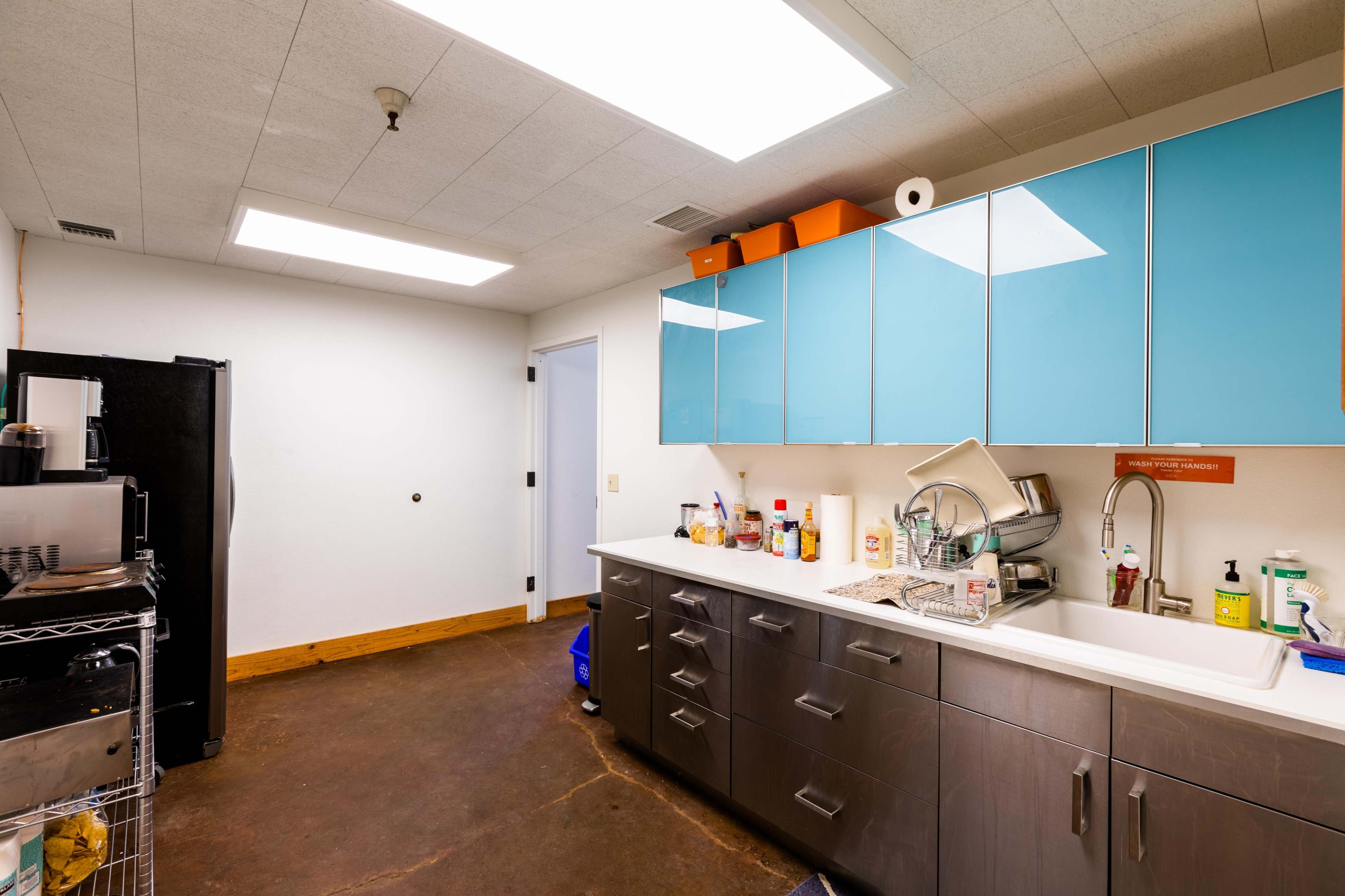 A kitchen with blue cabinets, a stainless steel sink, and appliances, featuring a concrete floor and overhead lighting.