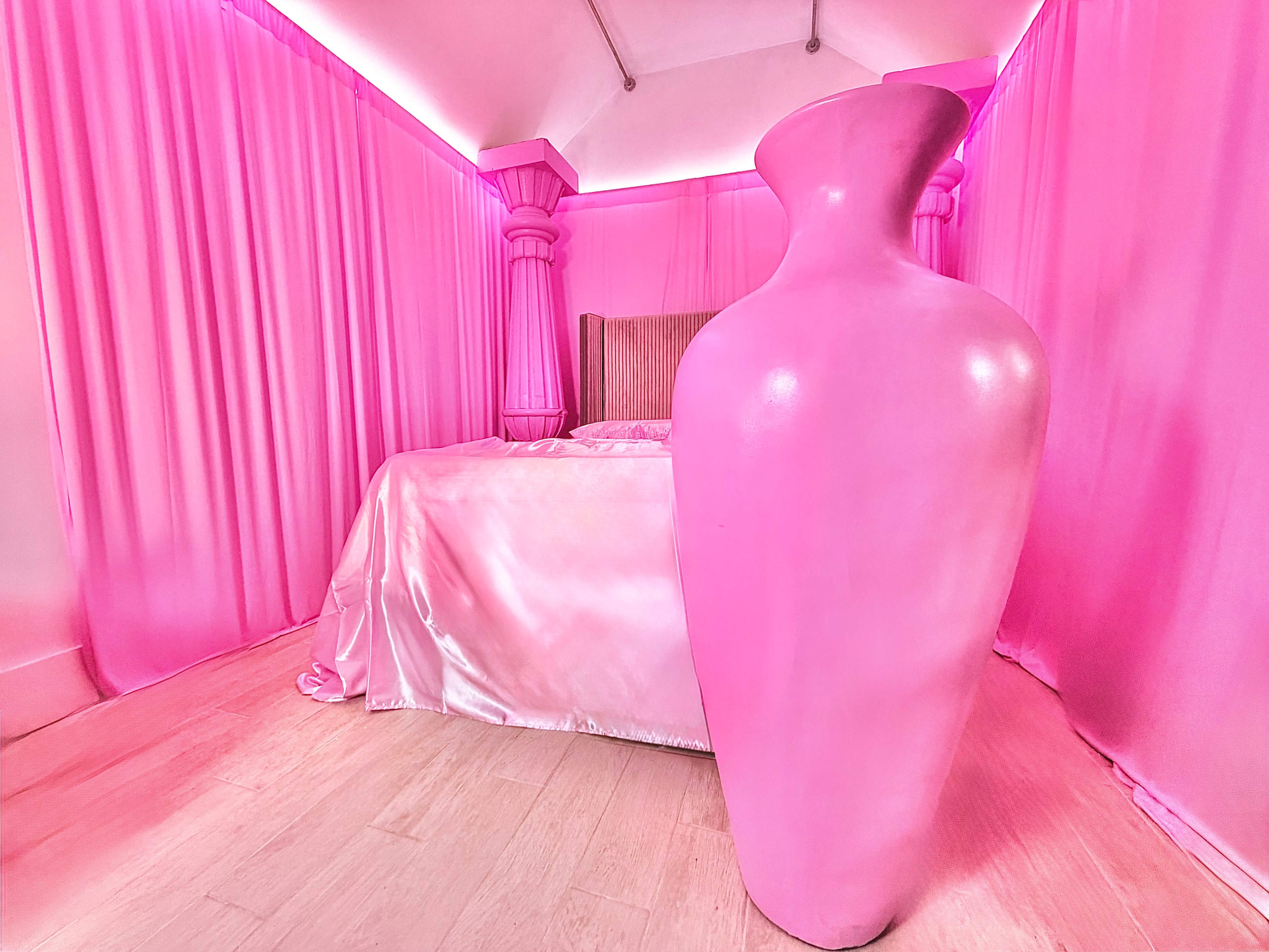 The image shows a room decorated in various shades of pink, featuring a large pink vase beside a bed draped in satin.