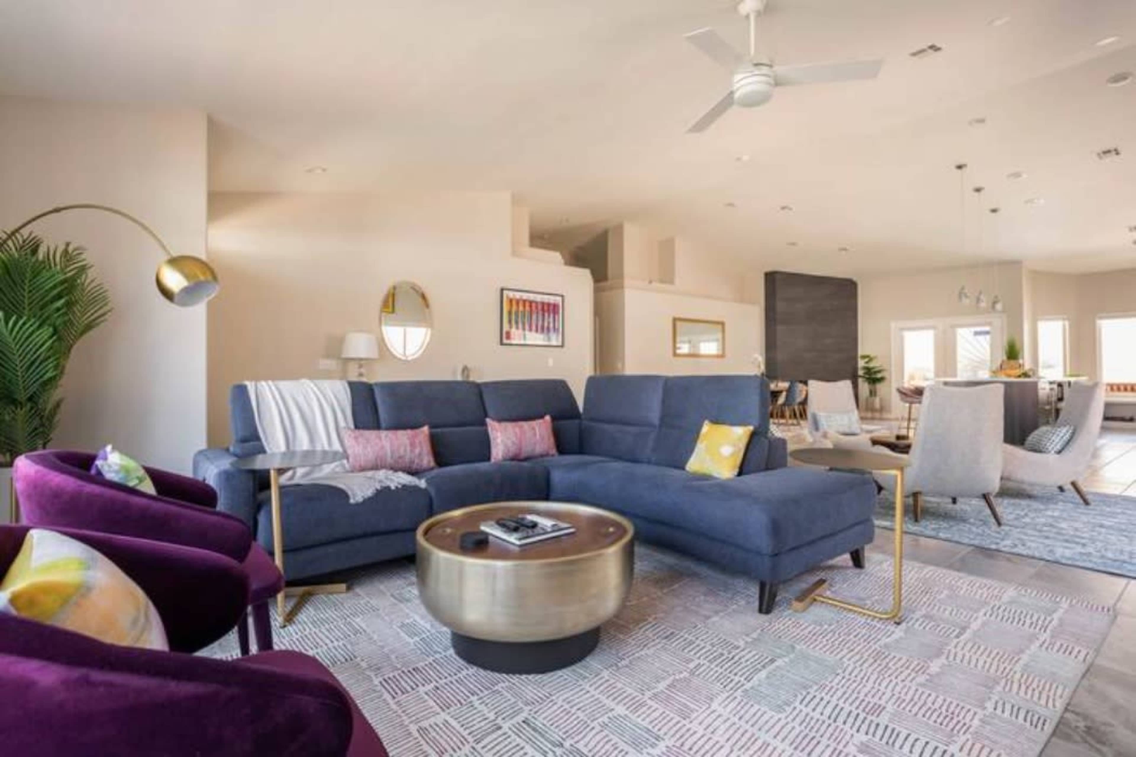 A modern living room features a blue sectional sofa, two purple armchairs, a round coffee table, and a patterned area rug, with large windows allowing natural light.