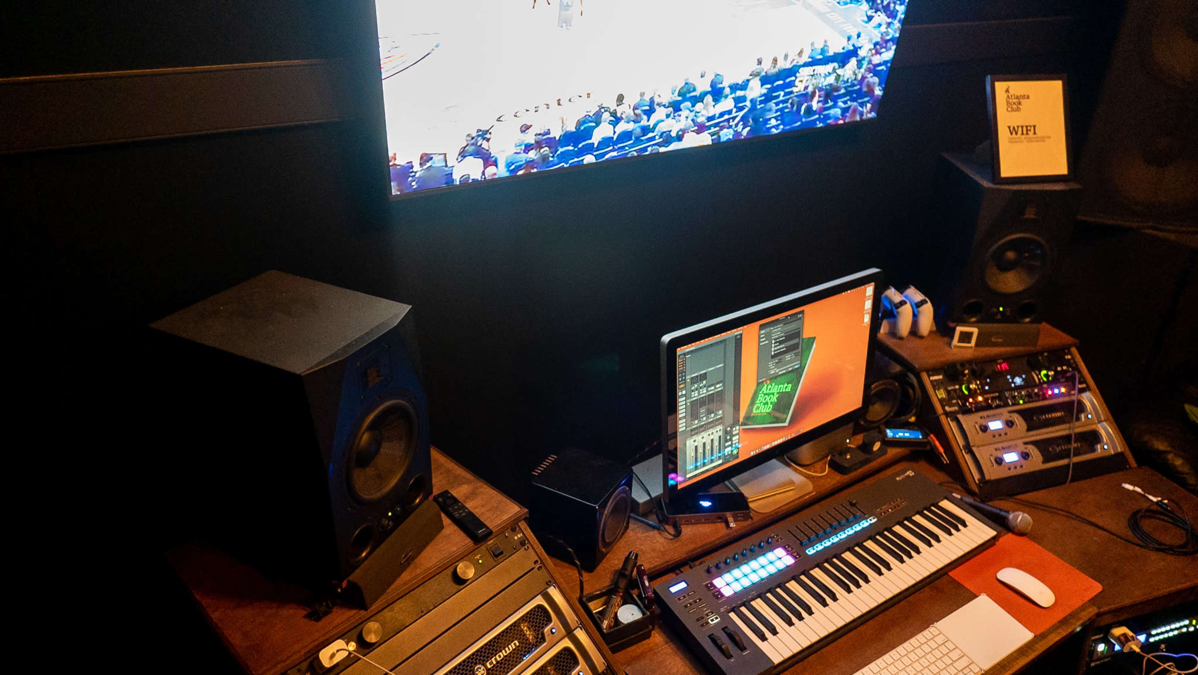 The image shows a music production setup featuring a keyboard, a computer display, and various audio equipment, with a sports game playing in the background on a large screen.