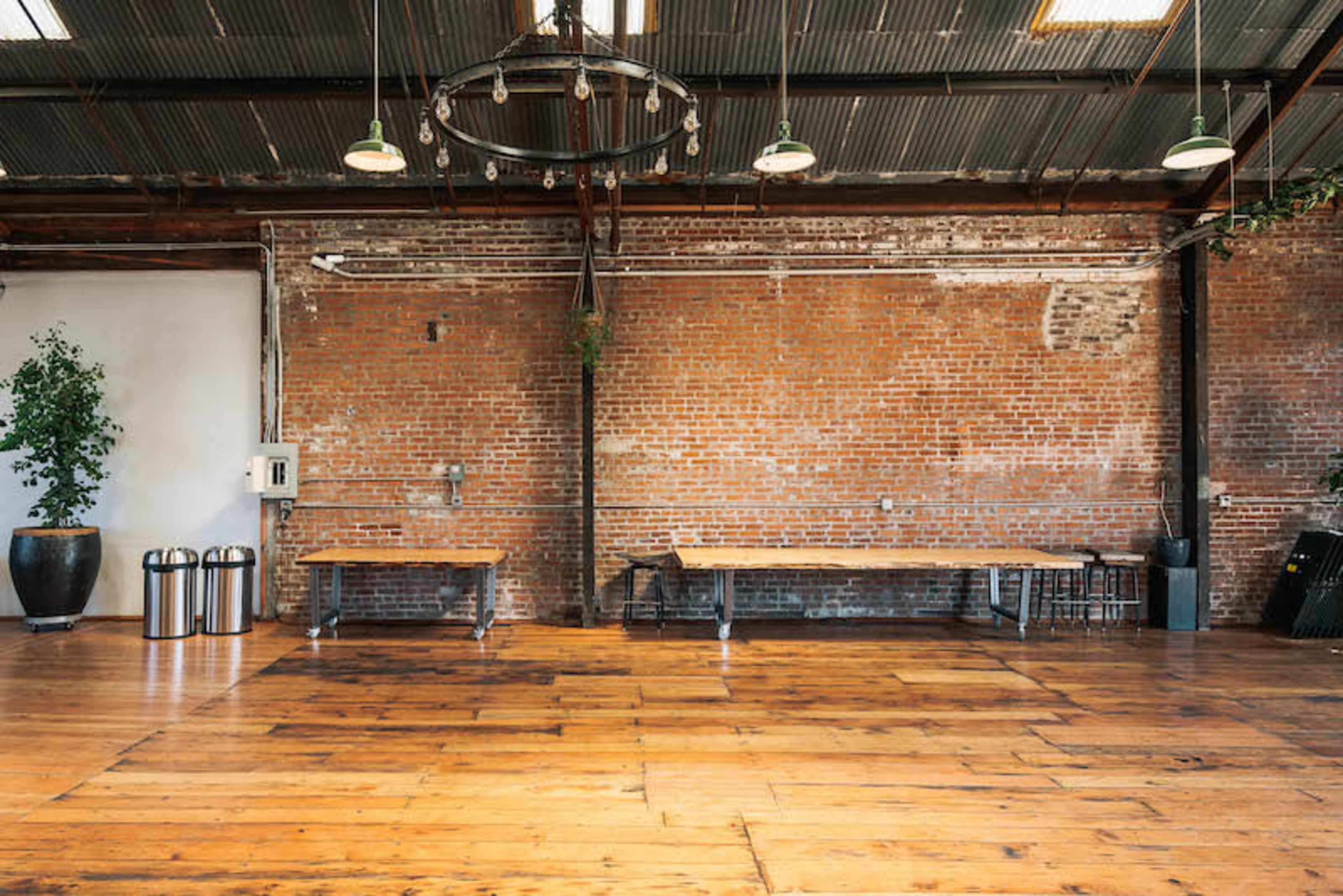 A spacious interior with exposed brick walls, wooden flooring, and simple metal tables arranged in the center.