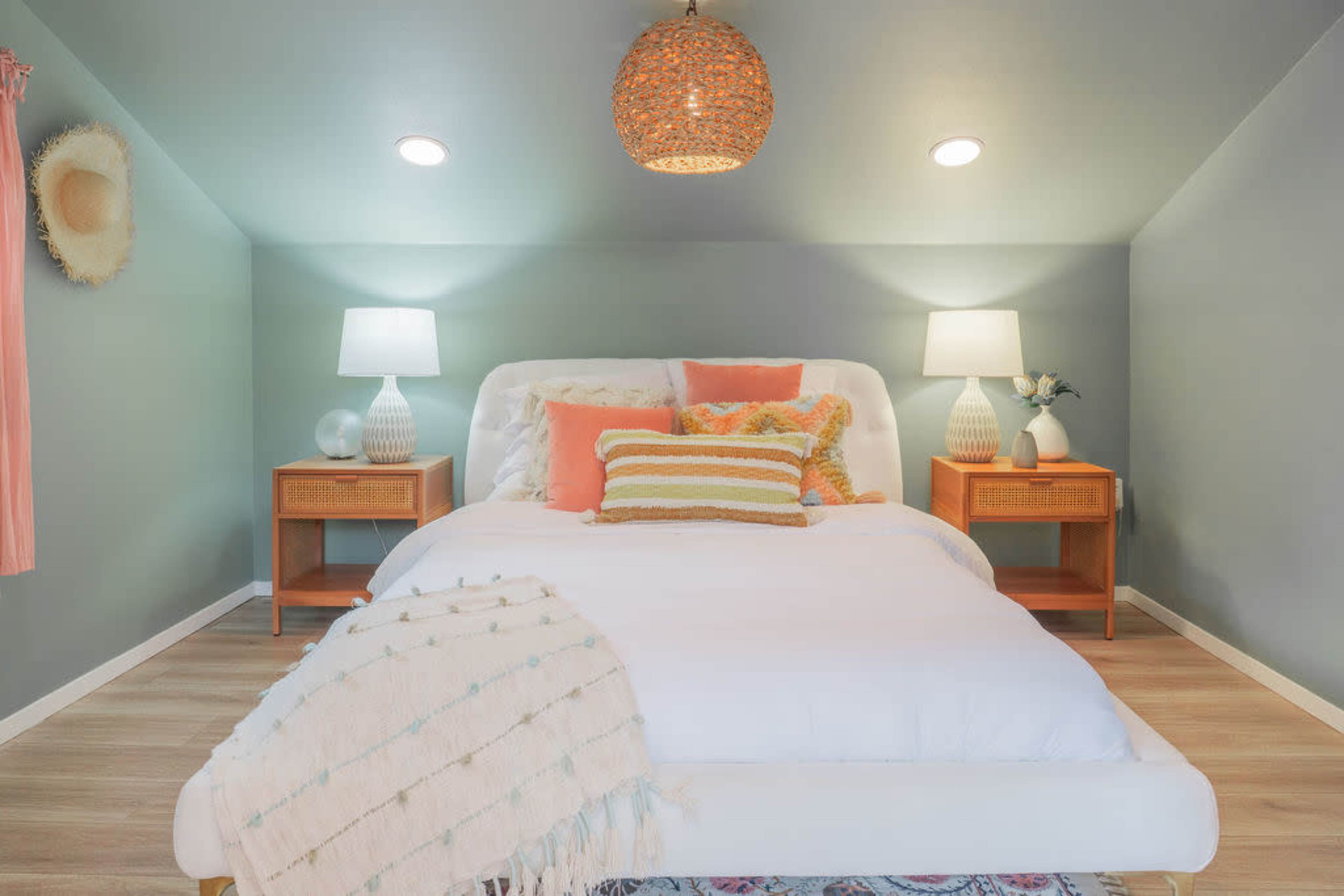 A neatly arranged bedroom features a white bed with colorful pillows, two matching bedside lamps, and textured decor against a soft green wall.