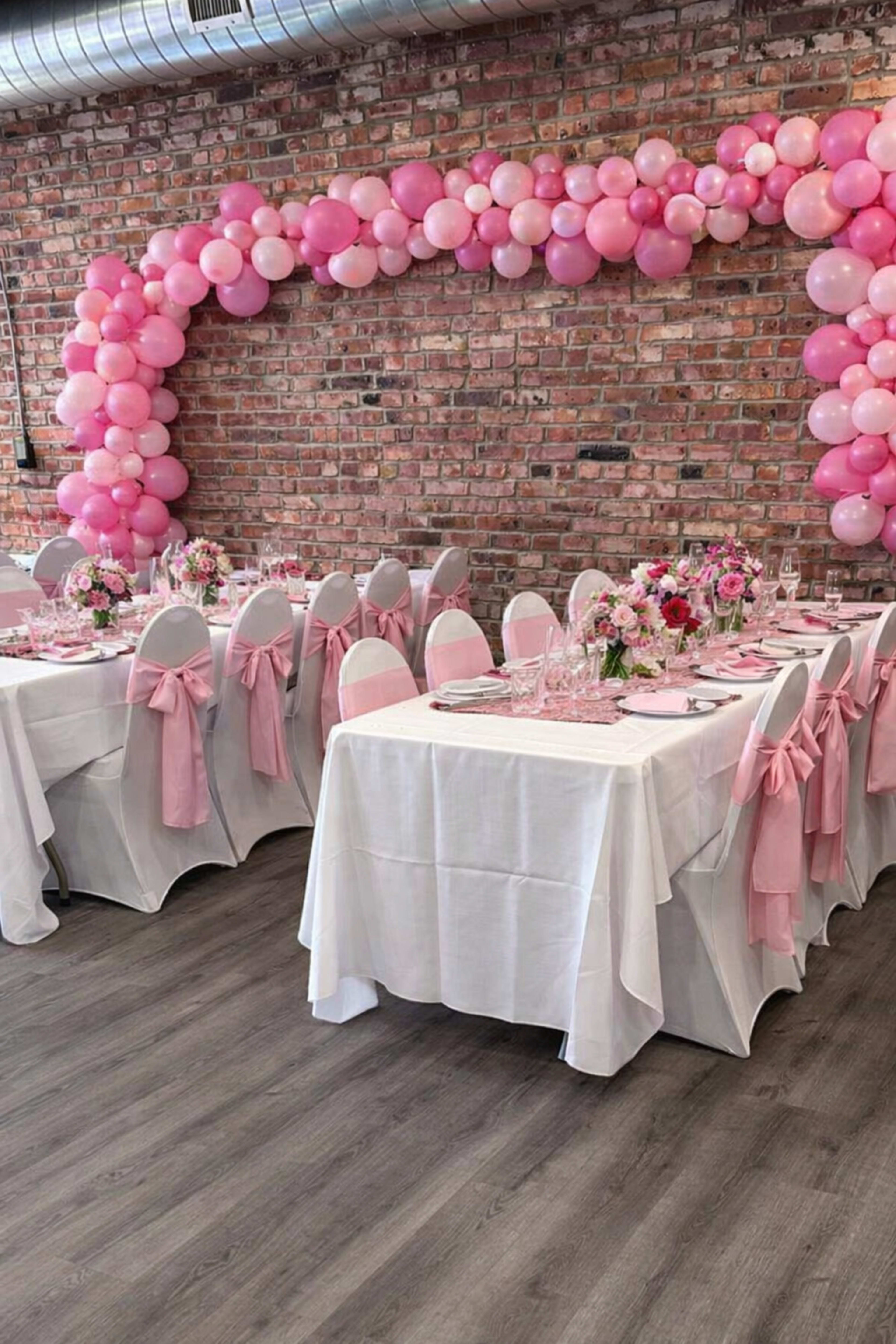 The image shows a decorated dining area with white tables covered in tablecloths, pink chair sashes, and a balloon arch made of pink balloons against a brick wall.