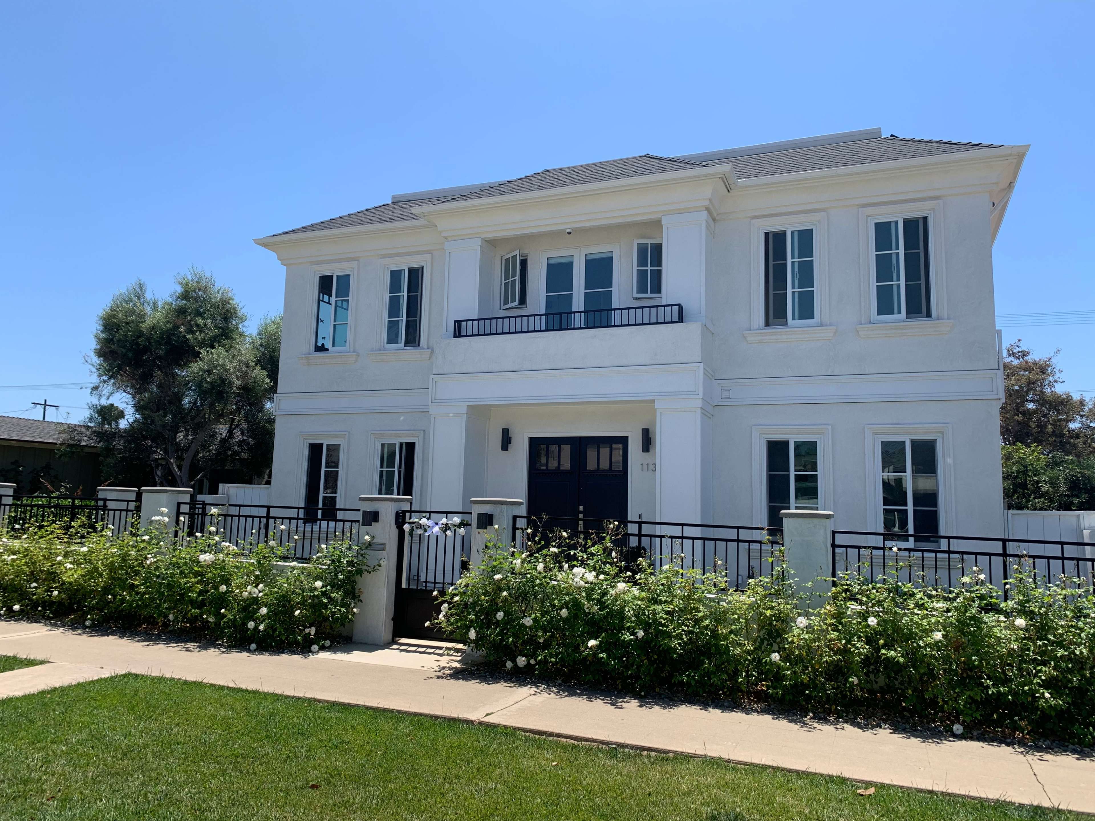 A two-story white house with black accents and a front balcony is surrounded by a well-maintained garden and lawn.