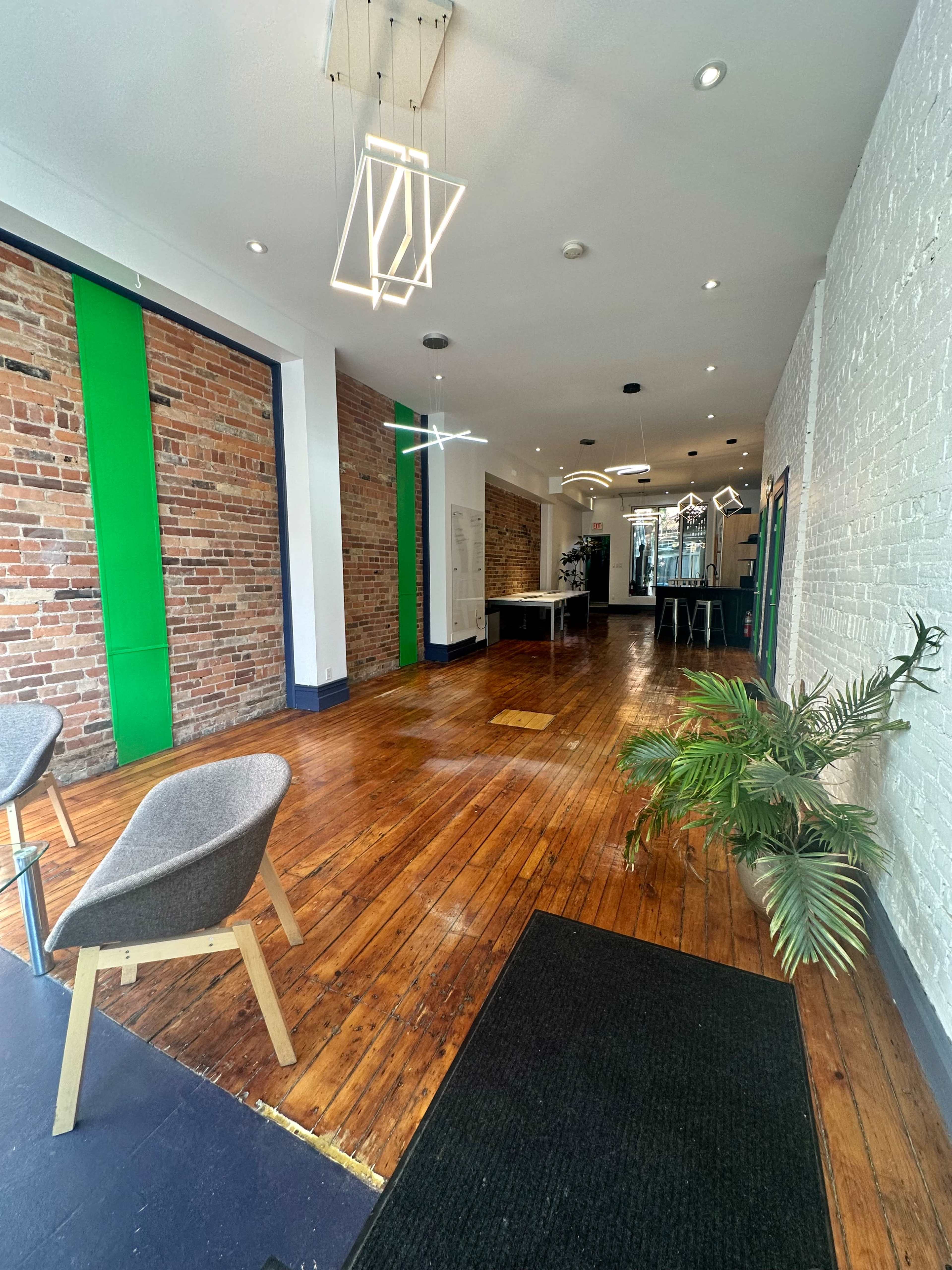 An empty interior space with wooden flooring, exposed brick walls, green accents, modern light fixtures, and a few pieces of furniture.