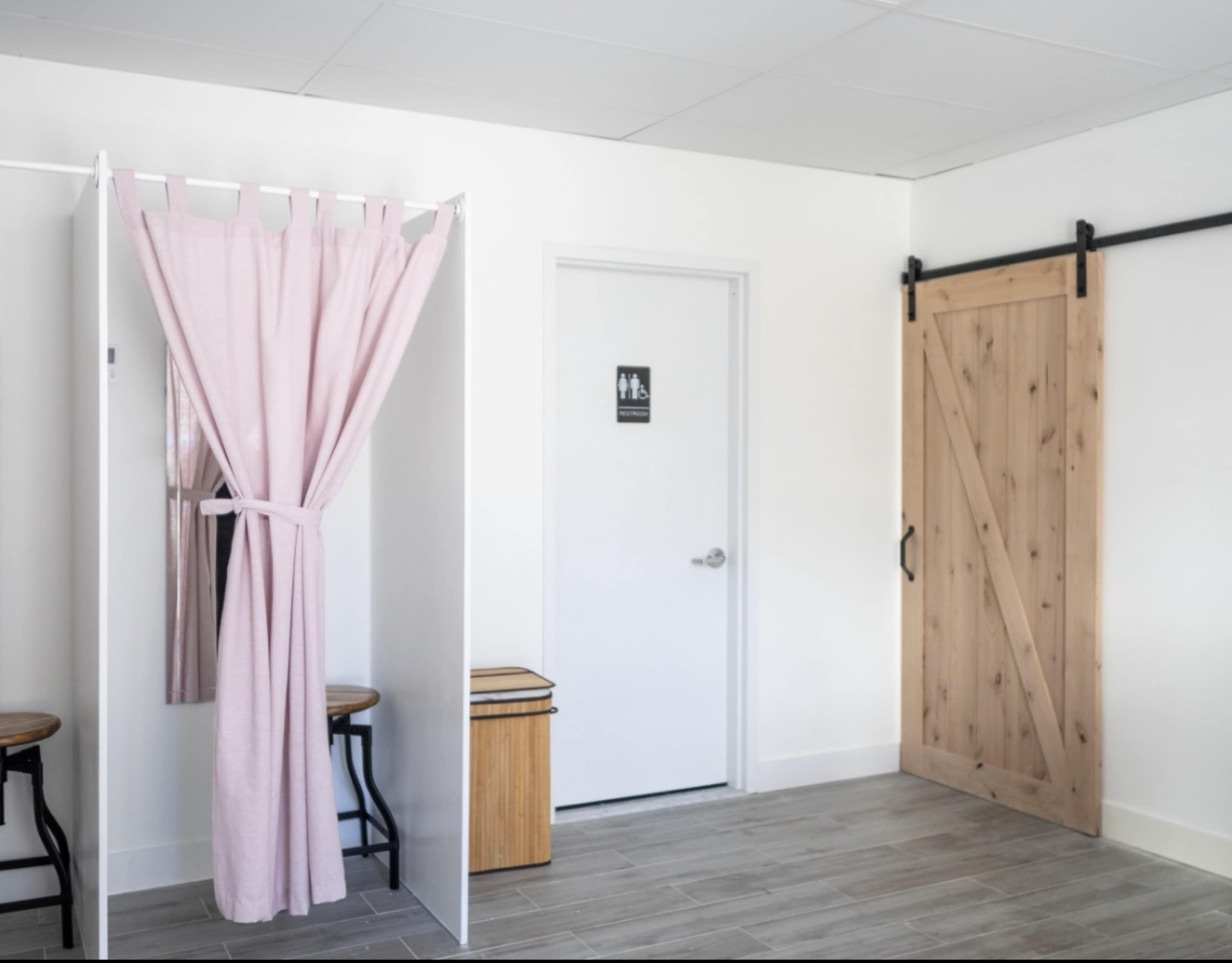 A bright, minimalist bathroom changing area with a pink curtain, a mirror, a door marked for restrooms, and a sliding wooden barn door.