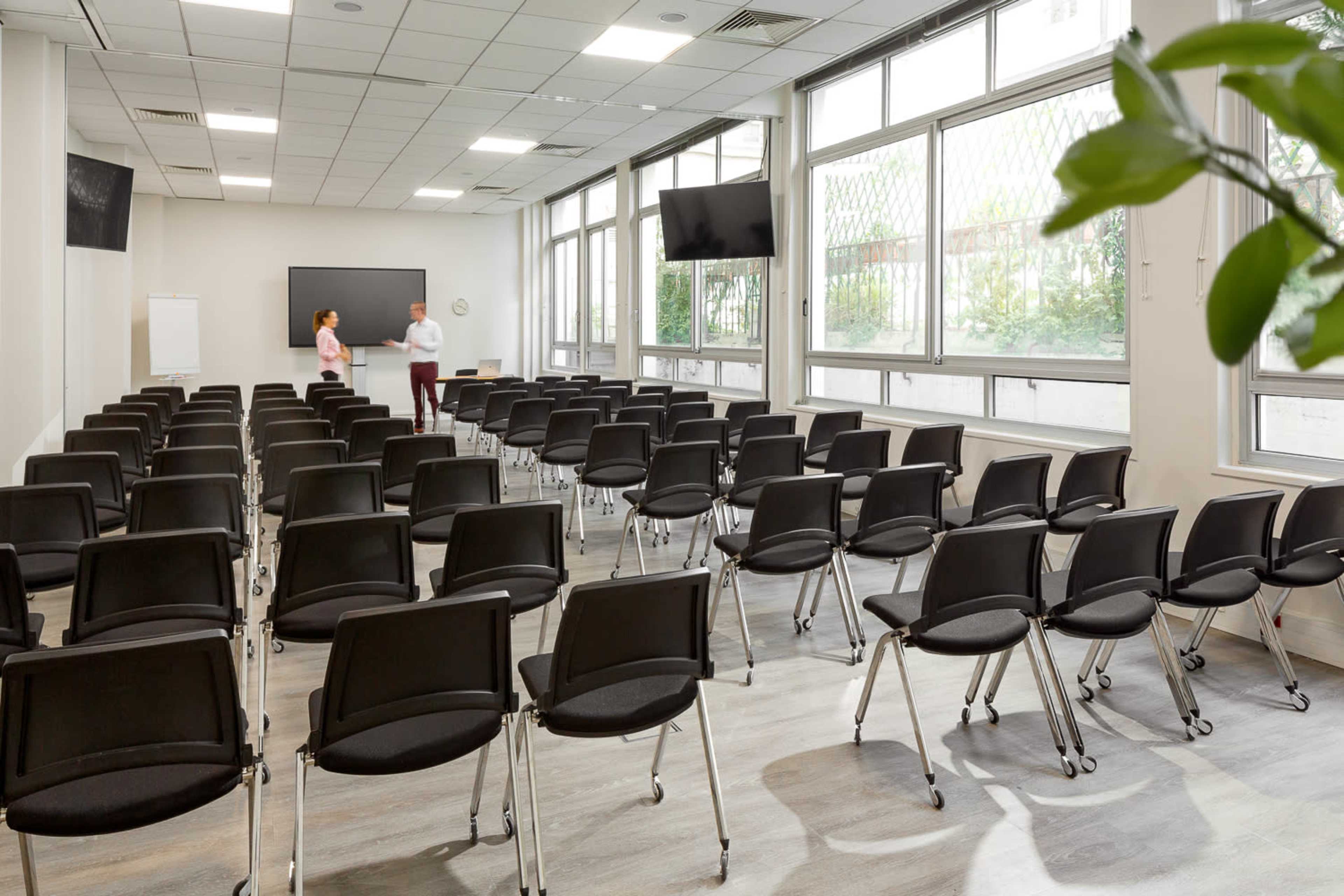 A modern conference room is set up with rows of empty black chairs facing a blank screen and two people conversing near the front.