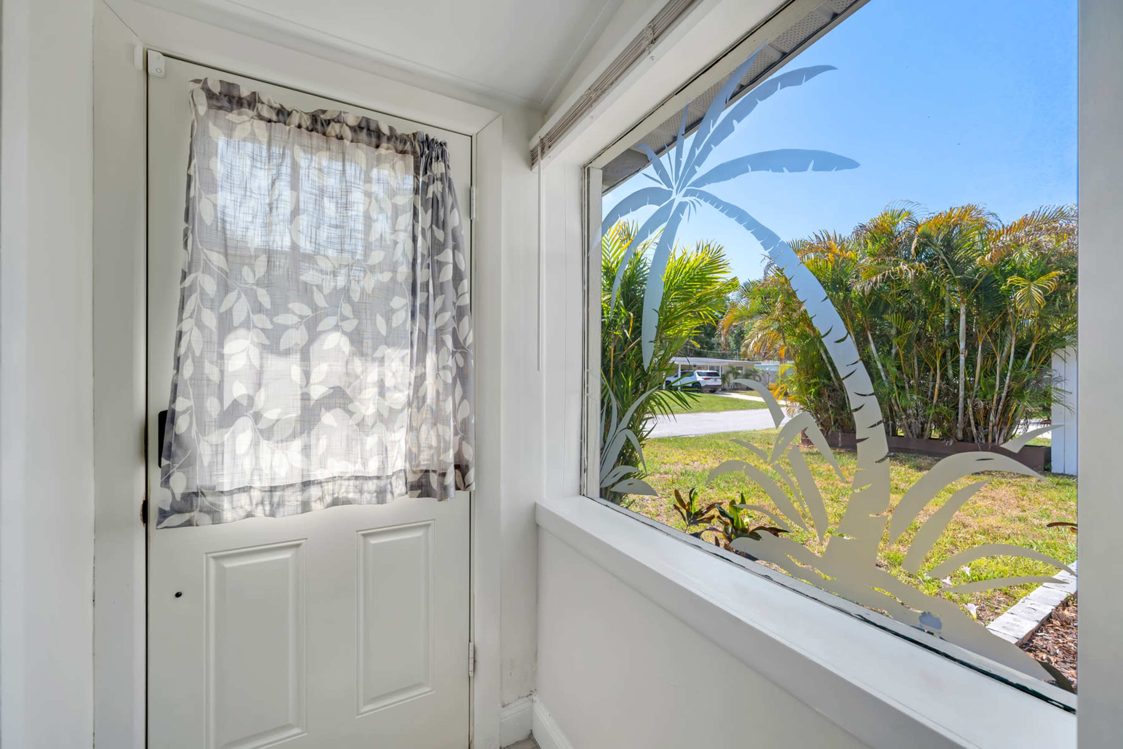 A white door with a sheer curtain is positioned next to a large window featuring a palm tree design, overlooking a landscaped yard with palm trees.