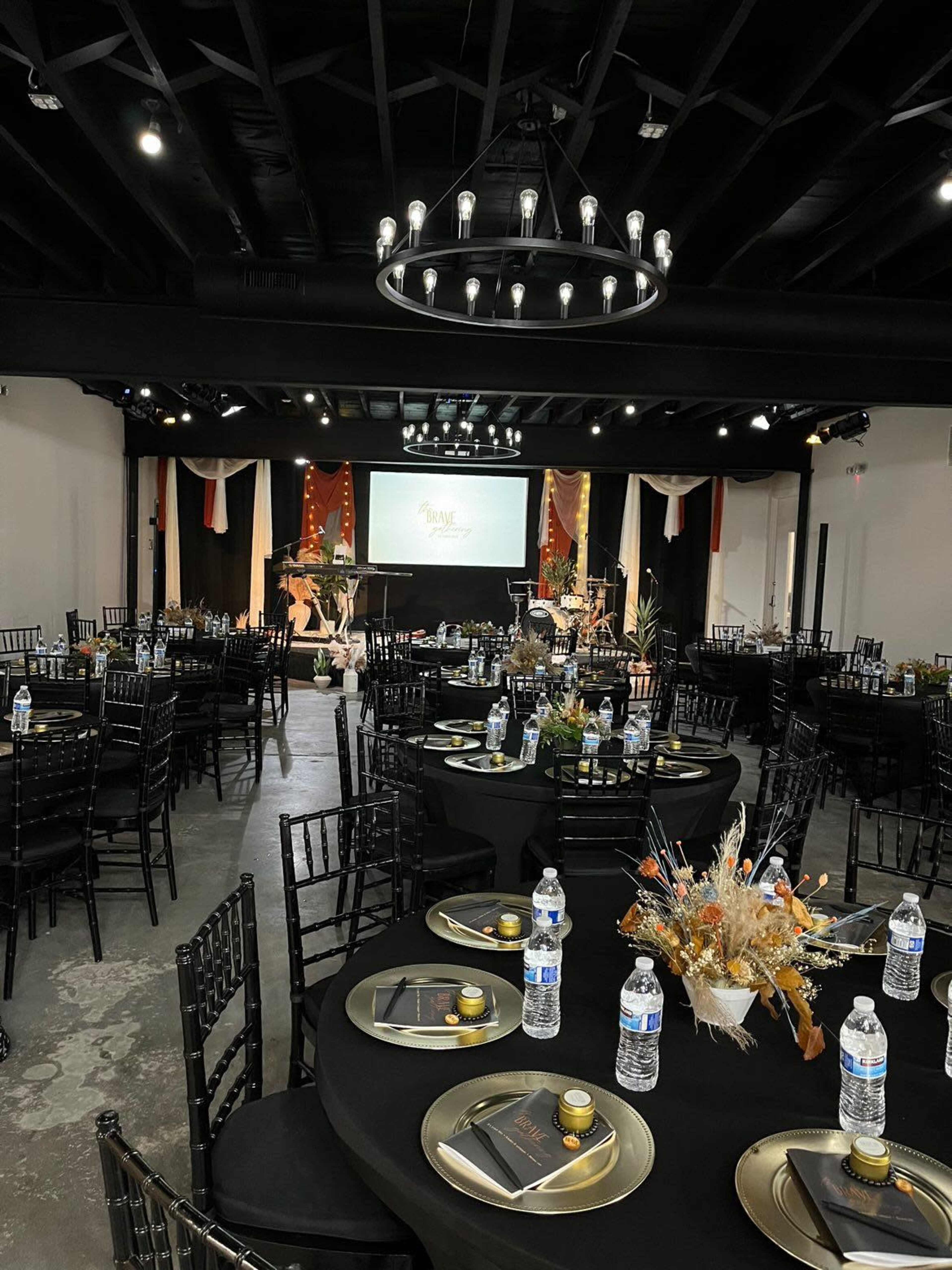 A banquet hall set up with round tables adorned with black tablecloths, centerpieces, and water bottles, under a chandelier and projection screen.
