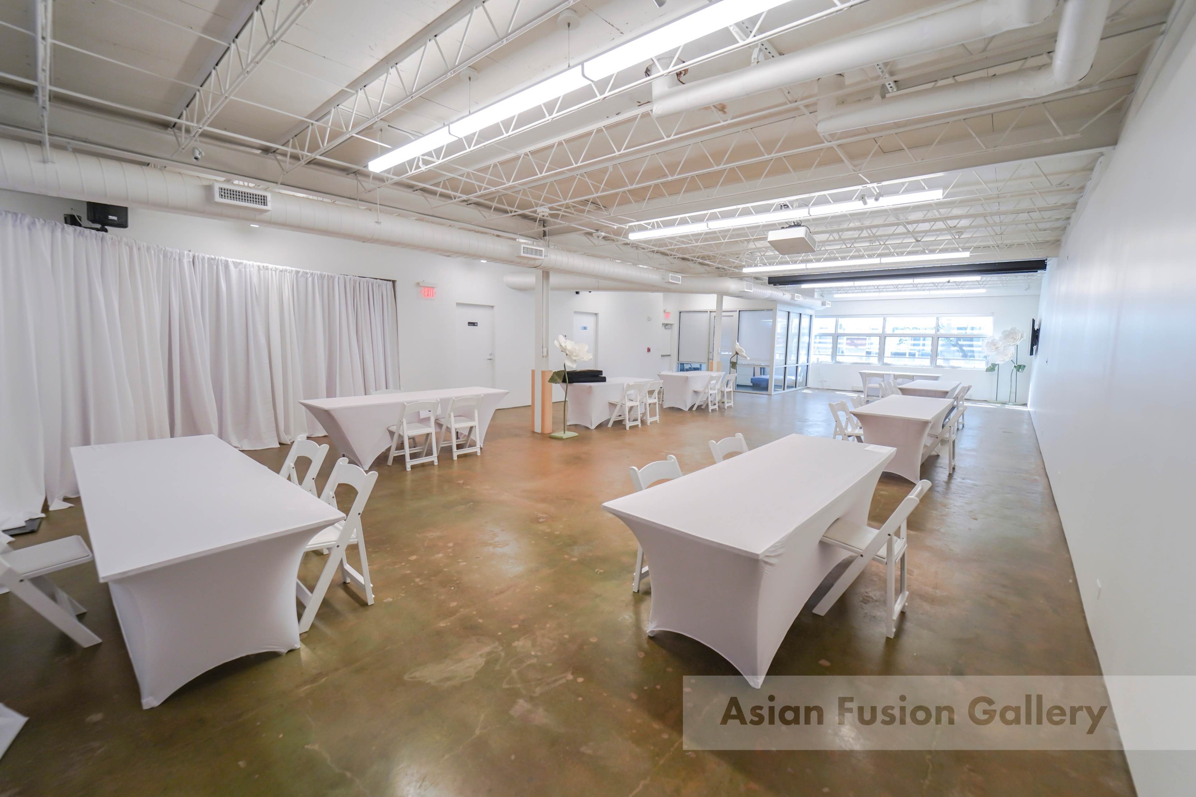A spacious gallery with multiple white table setups and a large window allowing natural light to enter.