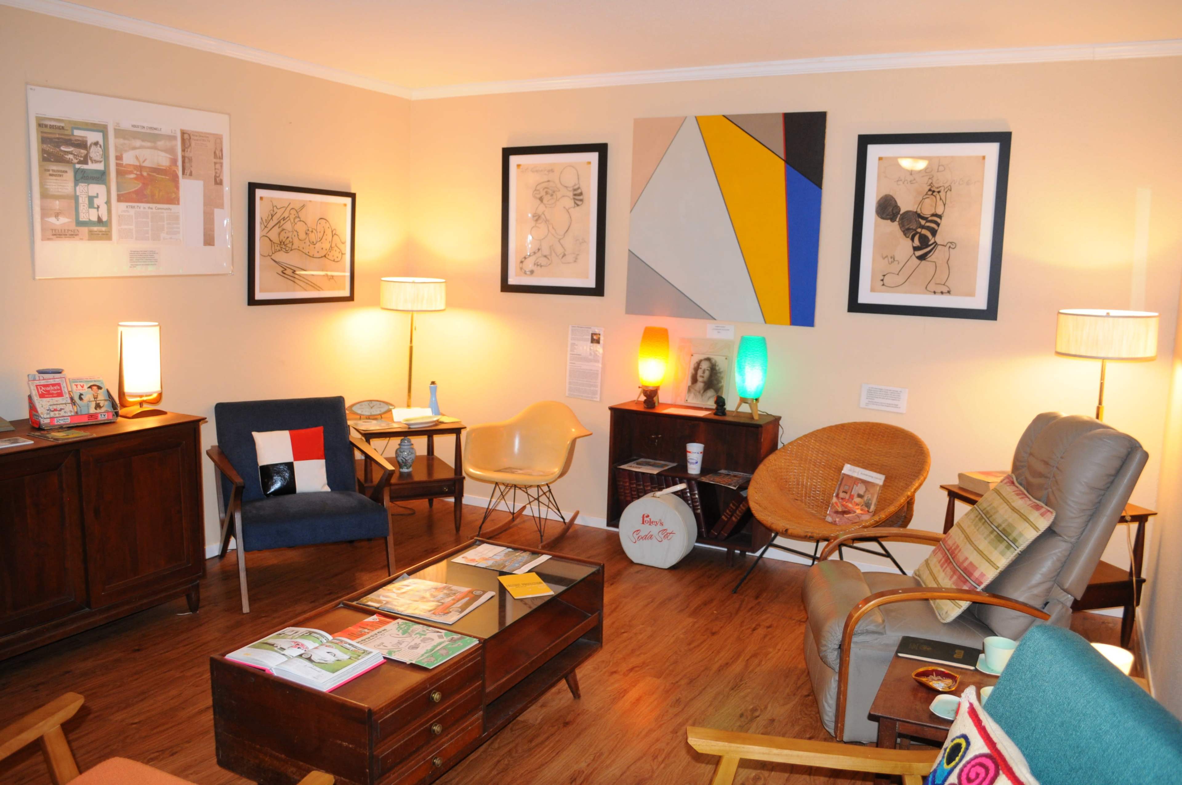 The image shows a cozy living room with various seating arrangements, colorful artwork on the walls, and a wooden coffee table topped with magazines.