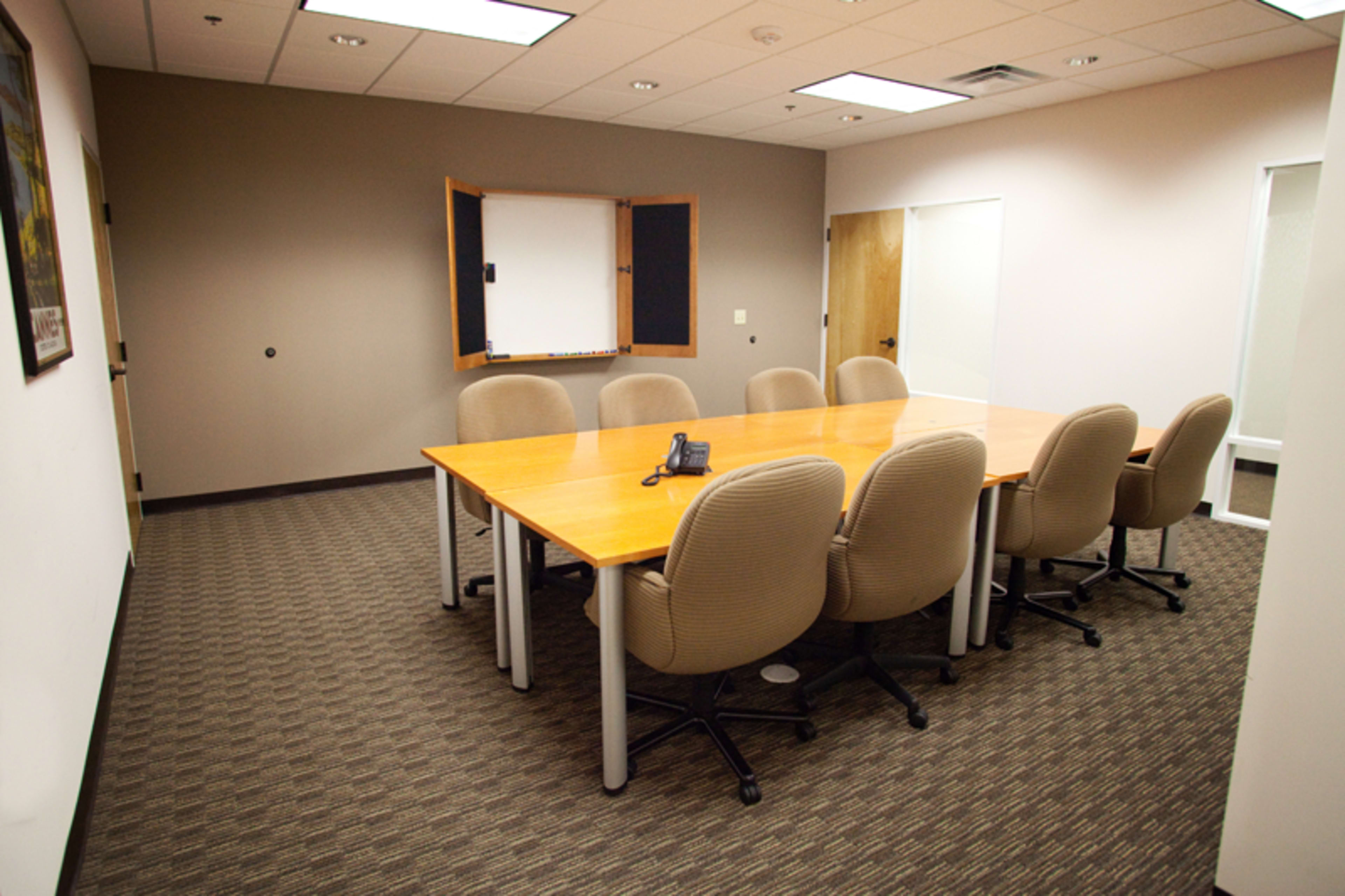 A conference room features a large wooden table surrounded by eight chairs, with a whiteboard mounted on the wall and a phone on the table.