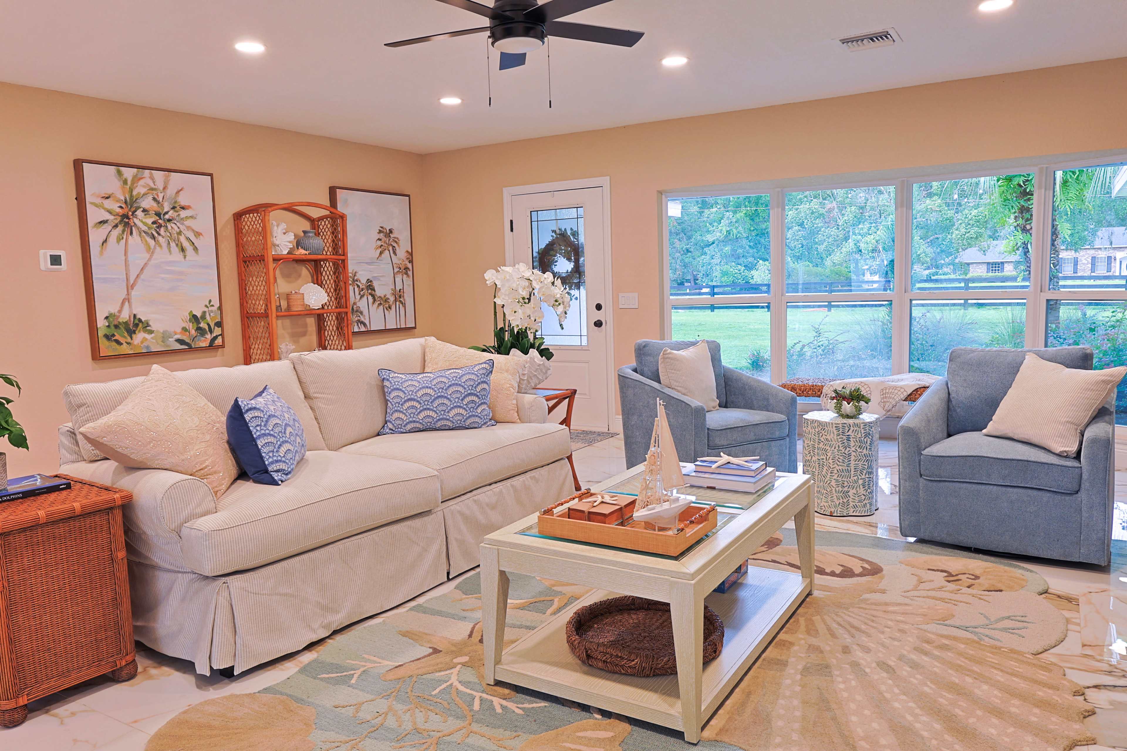 The image shows a light and airy living room featuring a beige sofa, two armchairs, a coffee table, and large windows overlooking a green outdoor space.