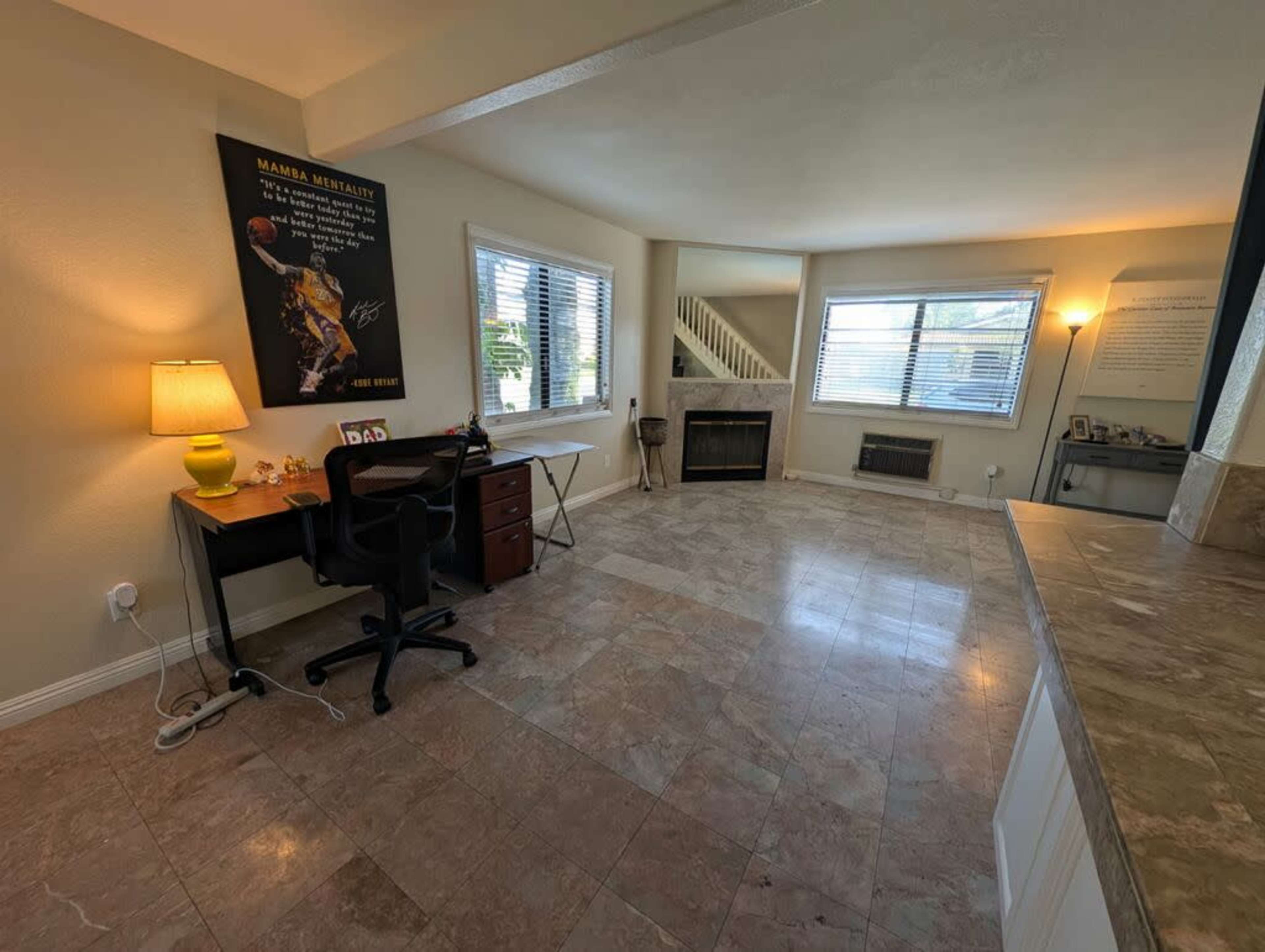 The image shows a light-filled living room with a desk, chair, and lamp, alongside a fireplace and large windows.