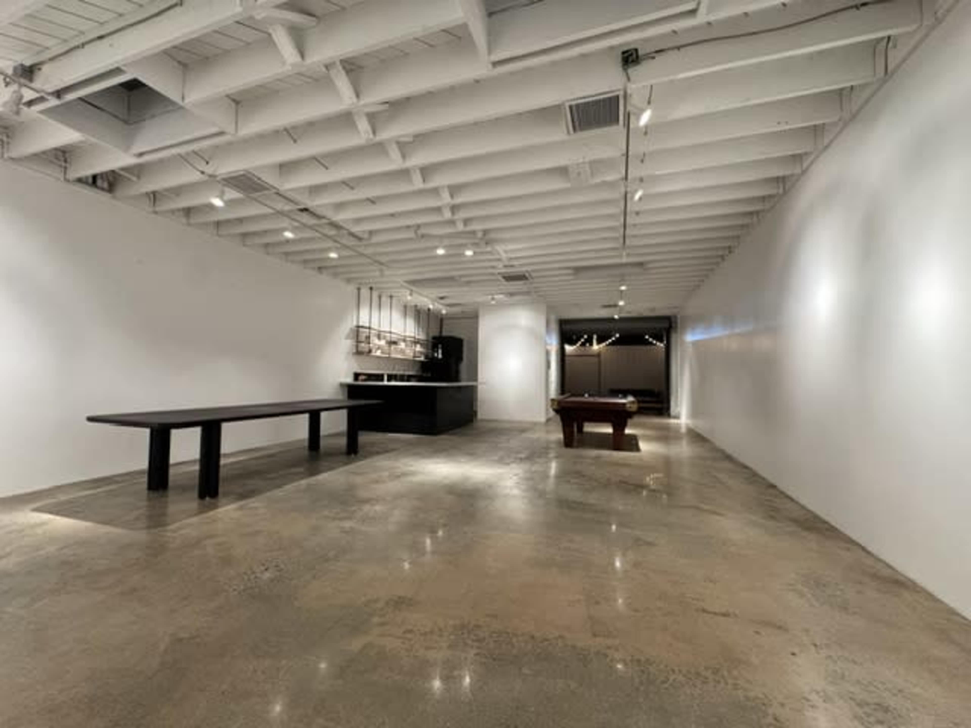 The image shows an expansive, sparsely furnished room with a polished concrete floor, a long black table, a pool table, and white walls.