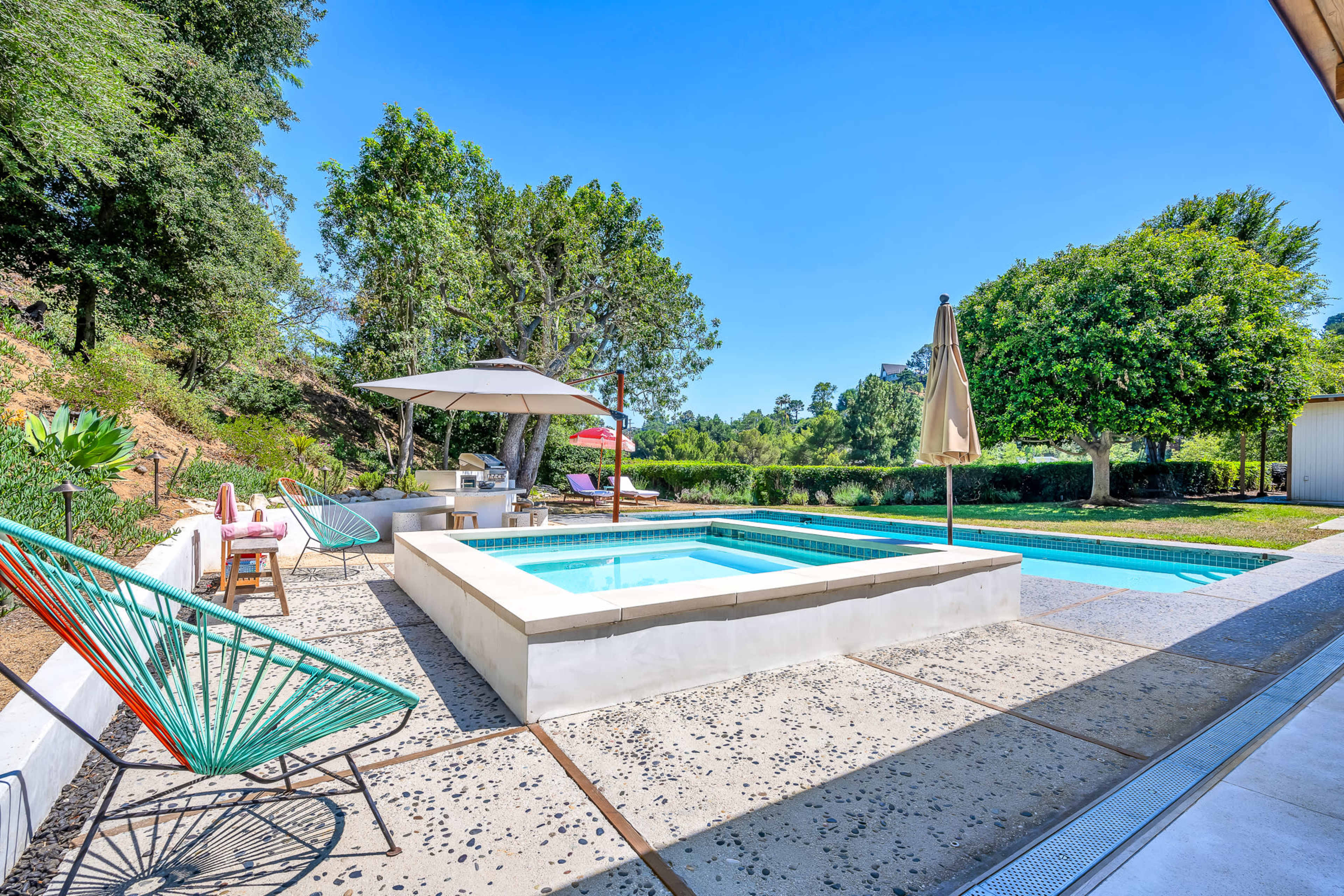 A spacious outdoor patio featuring a pool, a hot tub, lounge chairs, and a shaded sitting area surrounded by greenery.