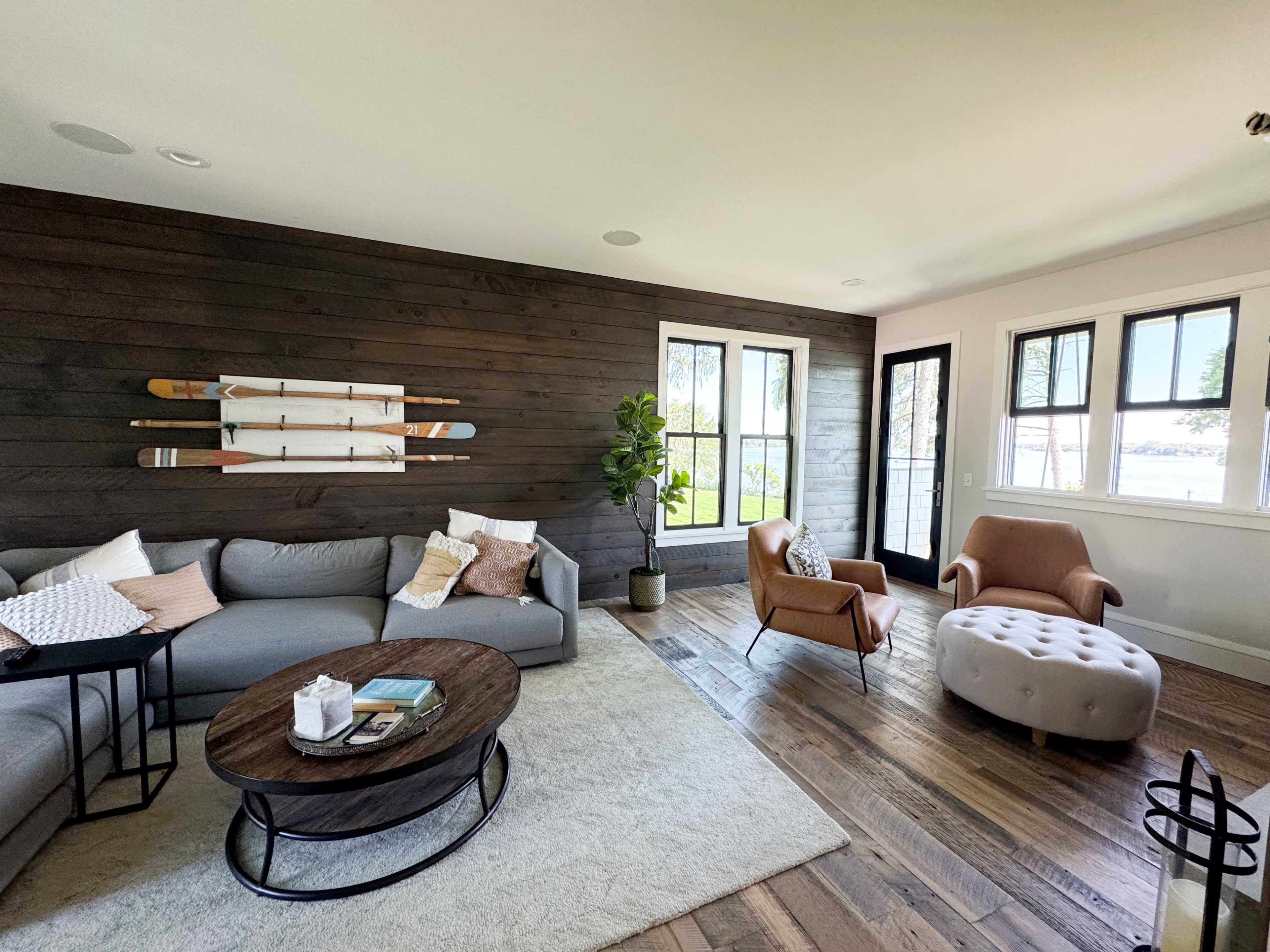 A modern living room features a gray sectional sofa, an accent chair, a round wooden coffee table, and a large window with a view of the outdoors.