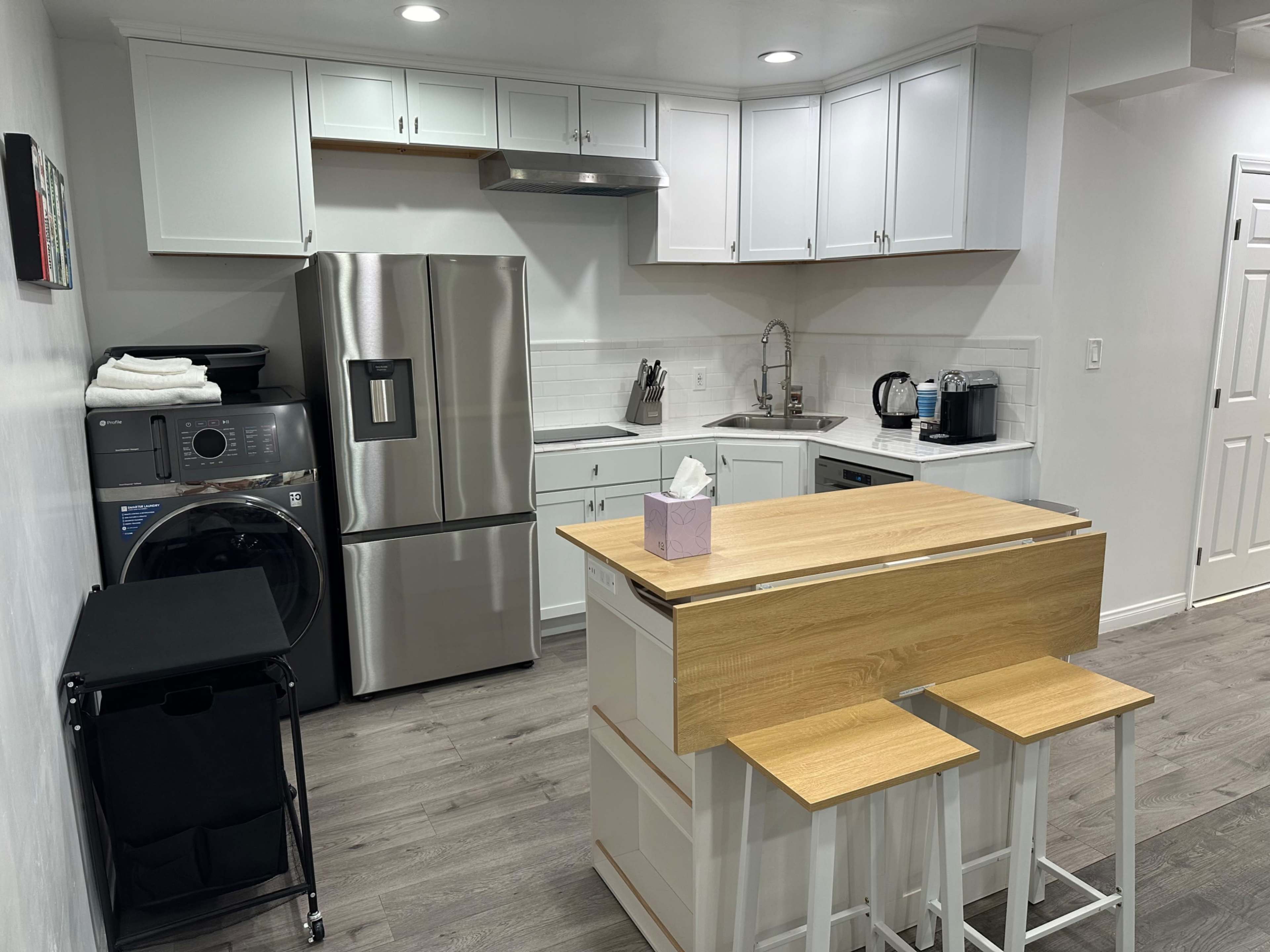 The image shows a modern kitchen featuring stainless steel appliances, a compact dining area with wooden stools, and a laundry area with a washing machine.