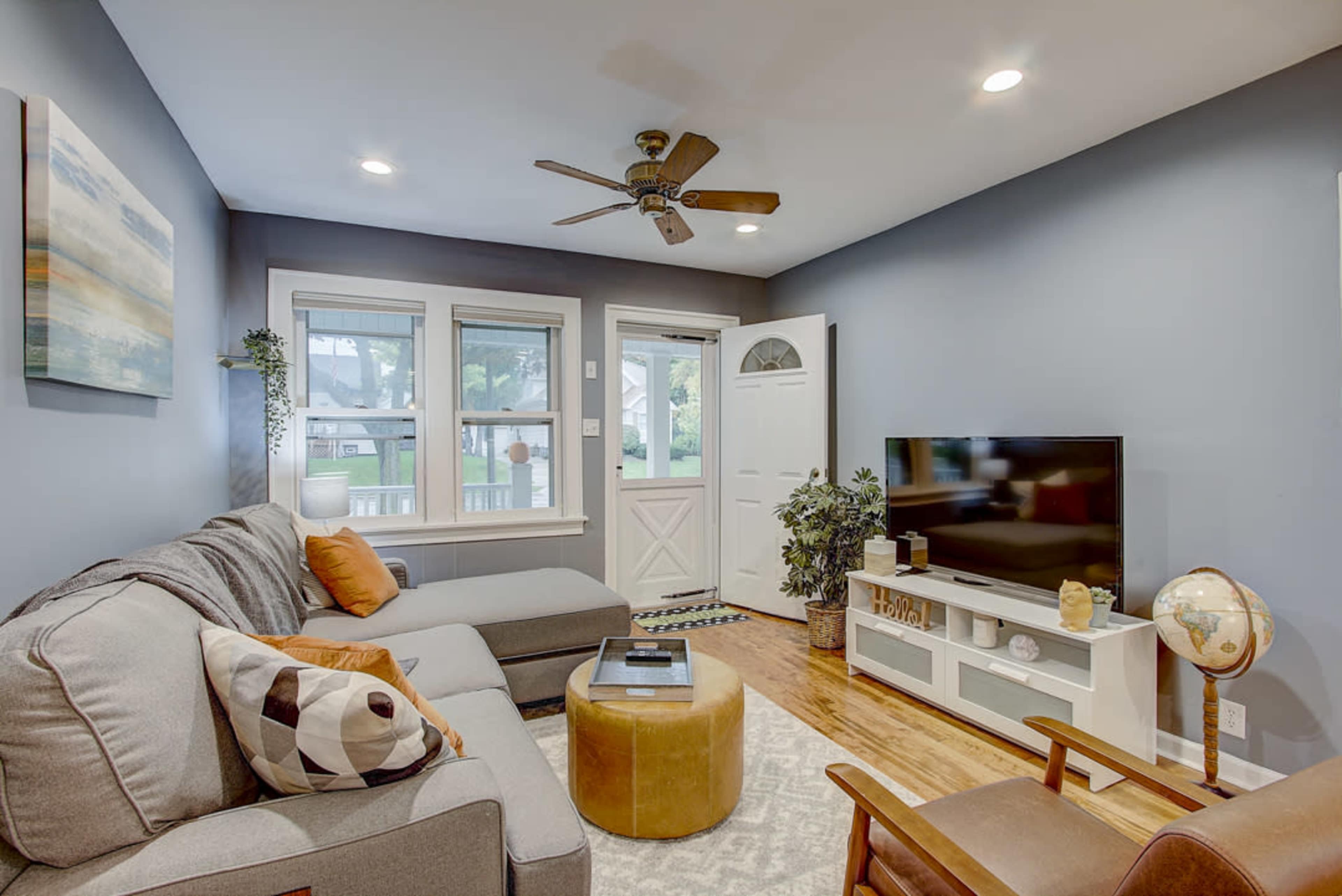 The living room features a gray sectional sofa, a television on a unit, a round ottoman, and large windows beside the front door.