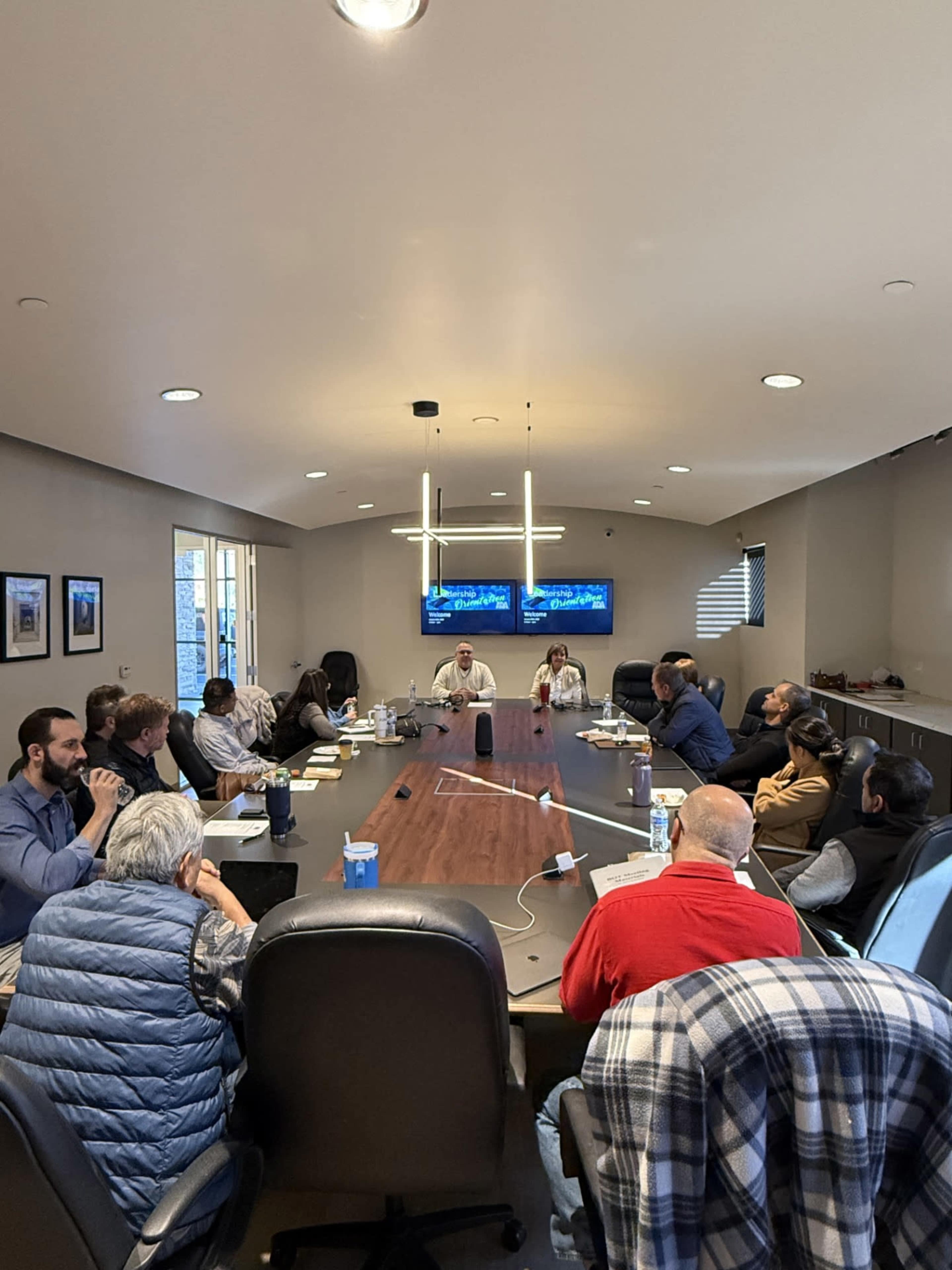 A group of professional individuals is seated around a large conference table in a well-lit meeting room, with multiple screens displaying information in the background.