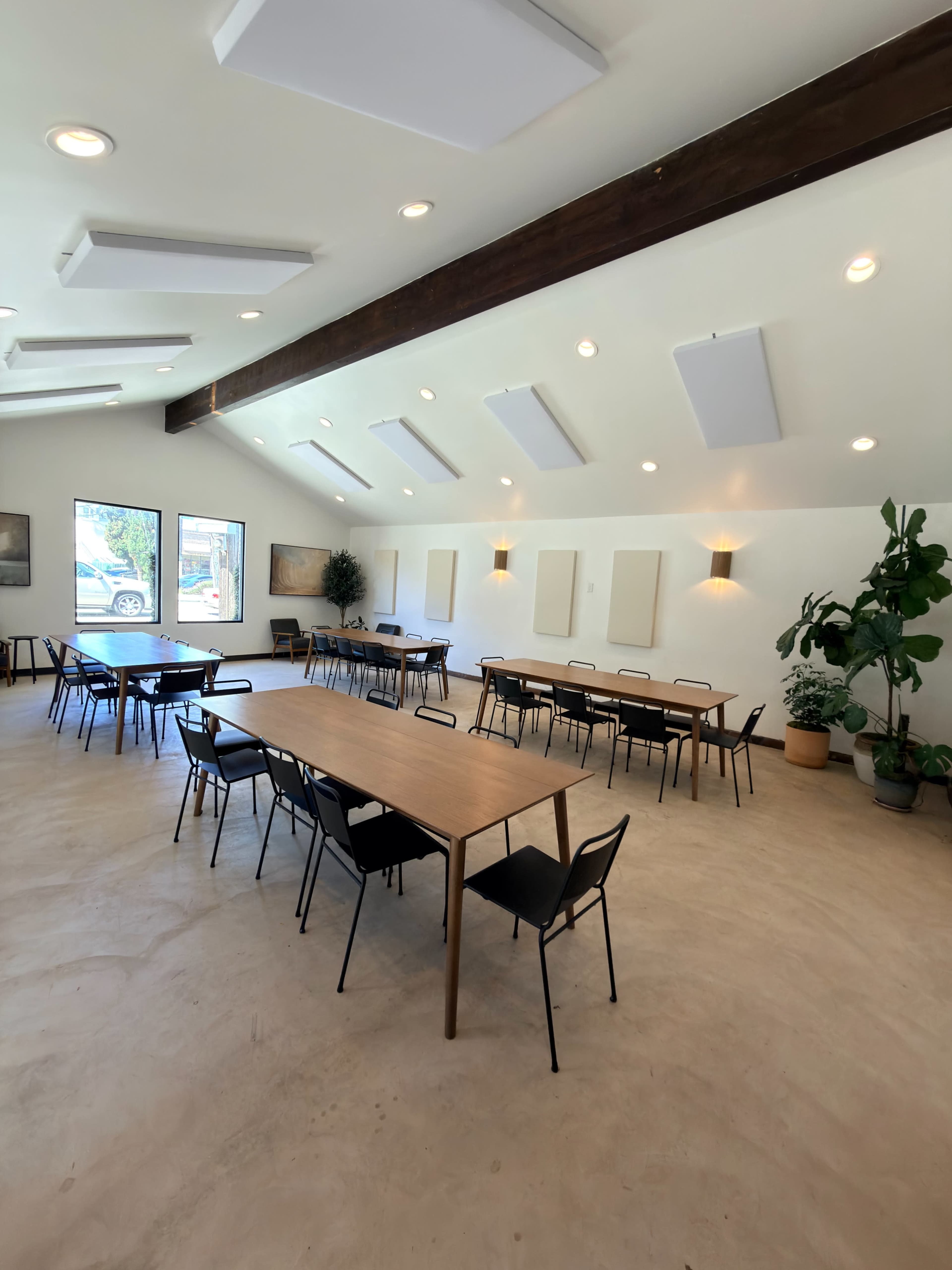 The image shows a spacious room set up for meetings or events, with multiple wooden tables and chairs arranged neatly along with decorative plants and wall-mounted sound panels.