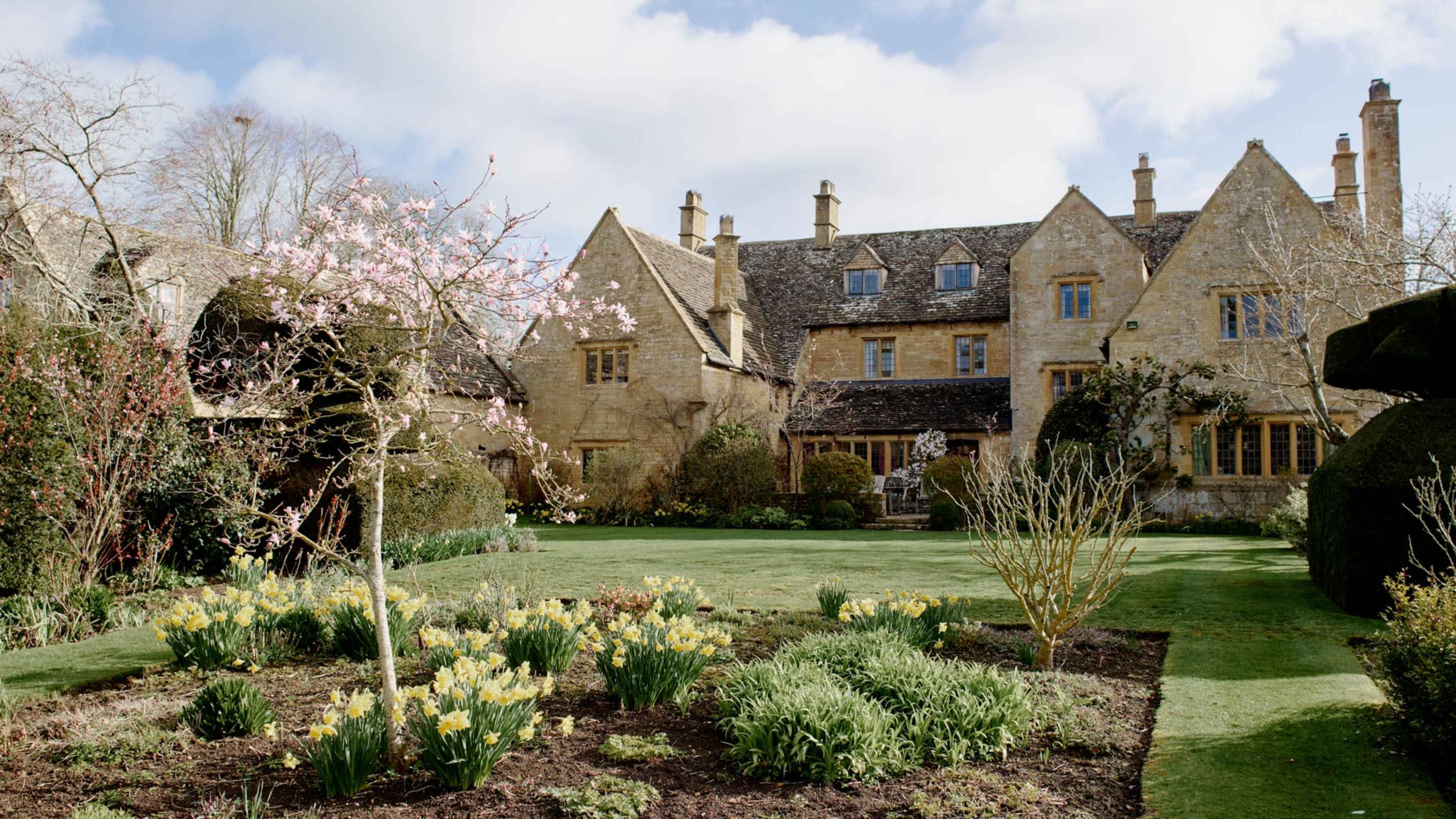 A well-manicured garden in front of a large stone house, featuring blooming trees and a variety of flowering plants.