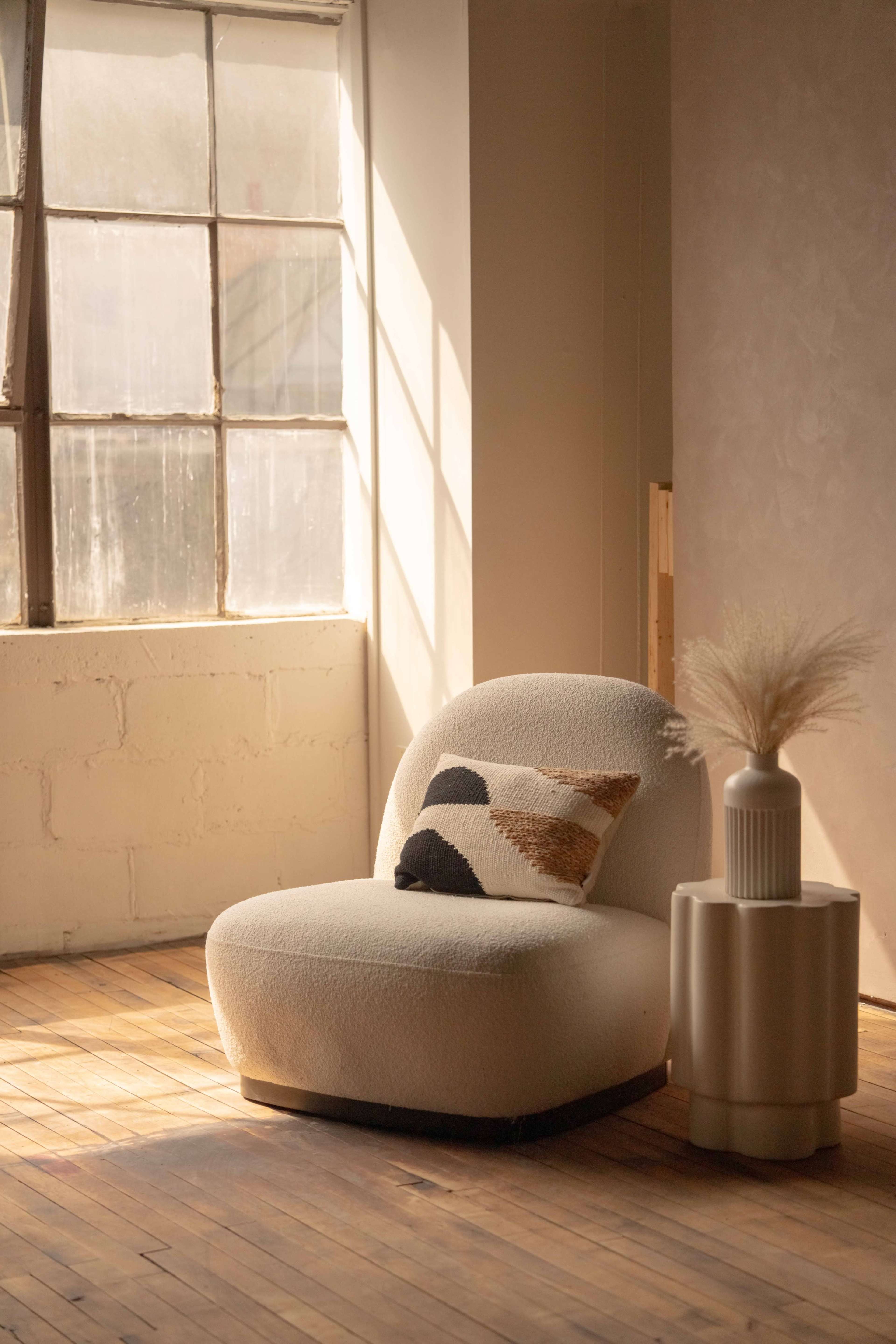 A curved, cream-colored chair with a decorative pillow sits beside a modern round side table in a sunlit room with large windows.