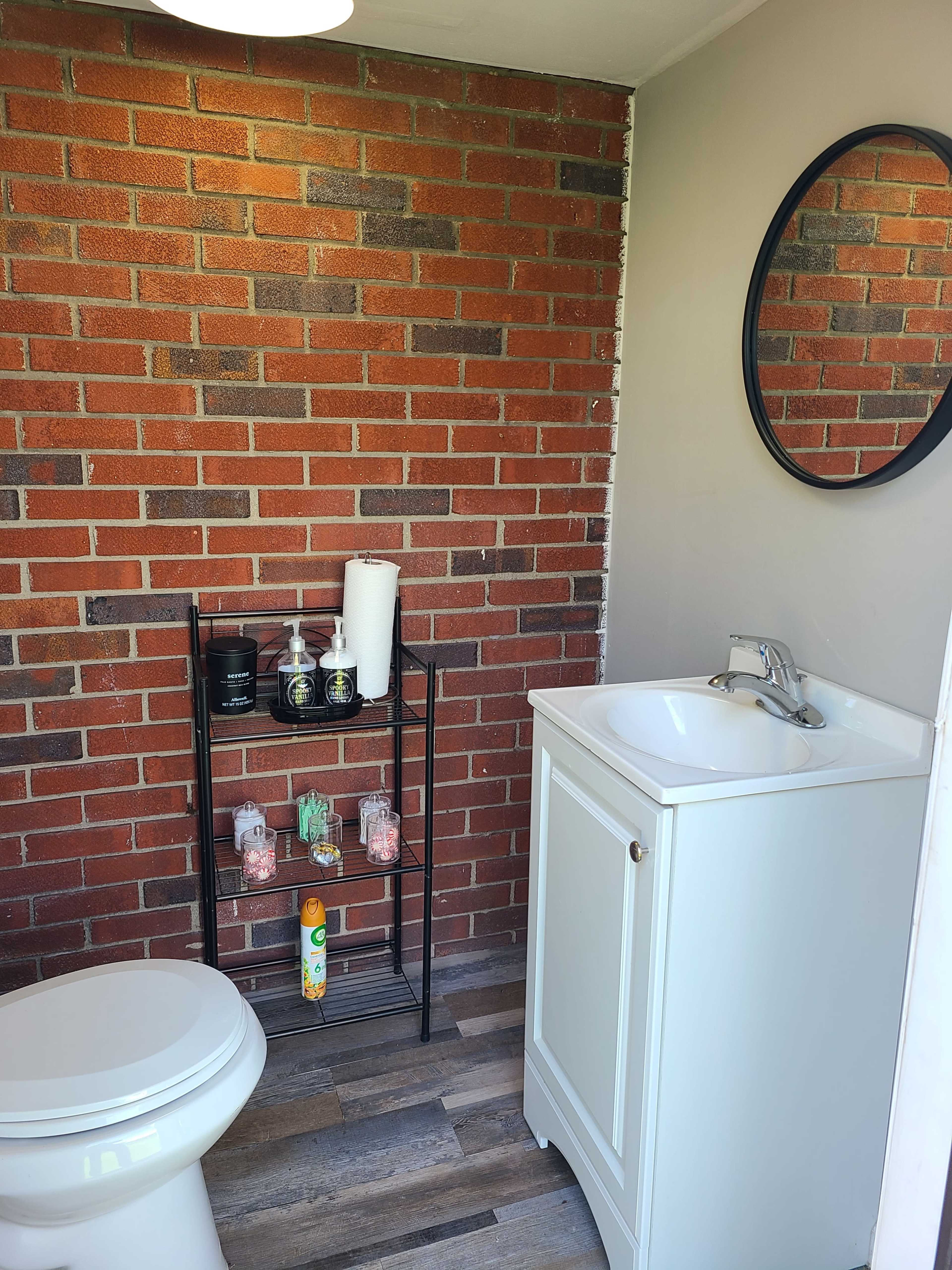 A bathroom featuring a white sink and toilet against a brick wall, with a black metal shelf displaying various toiletries.