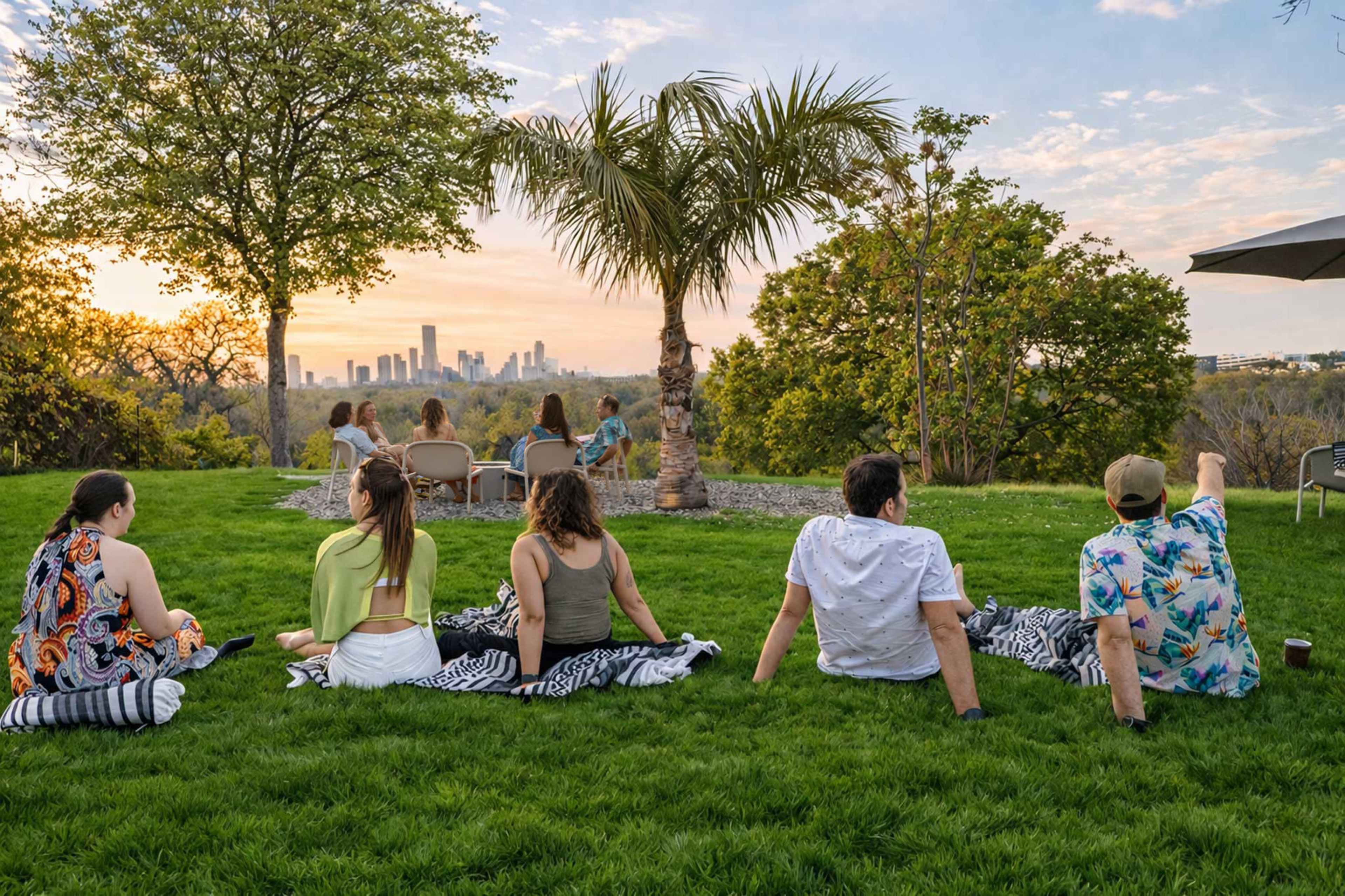 A group of six people sits on blankets in a grassy area, overlooking a city skyline at sunset.