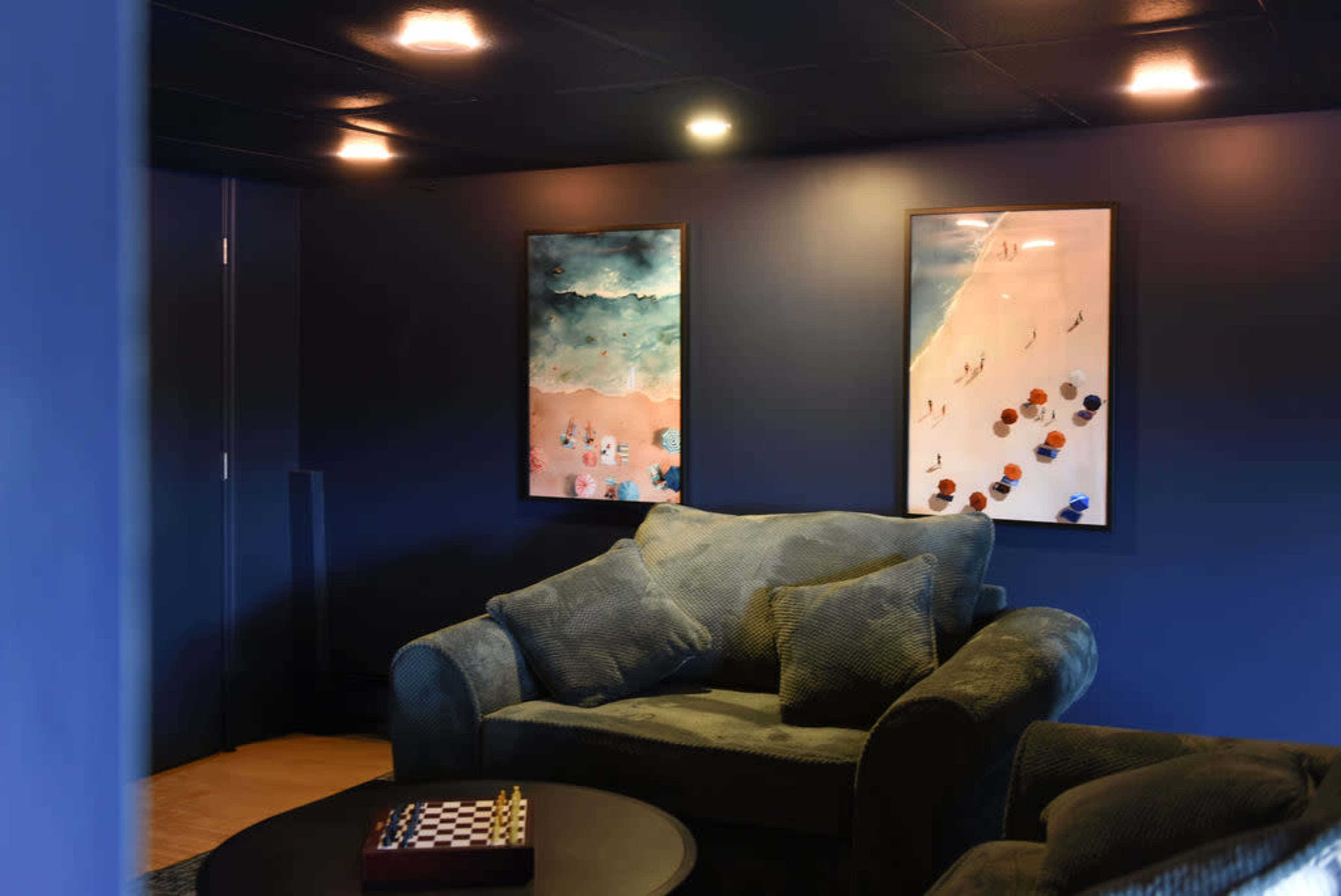 A cozy lounge area features two framed artworks on a dark blue wall, a plush sofa, and a low coffee table with a chess set.