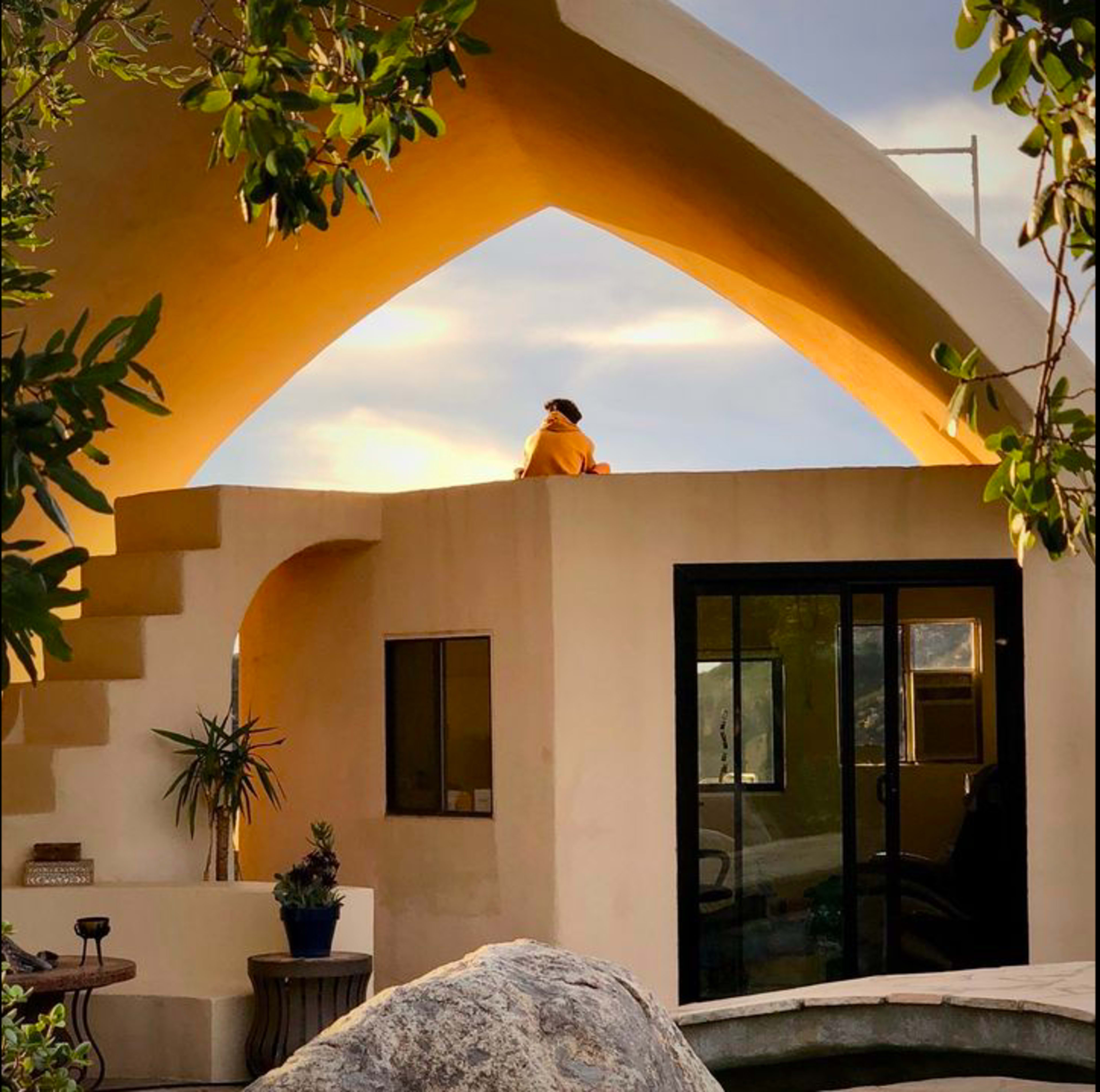 A person sits on the edge of a roof beneath a curved architectural structure during sunset.