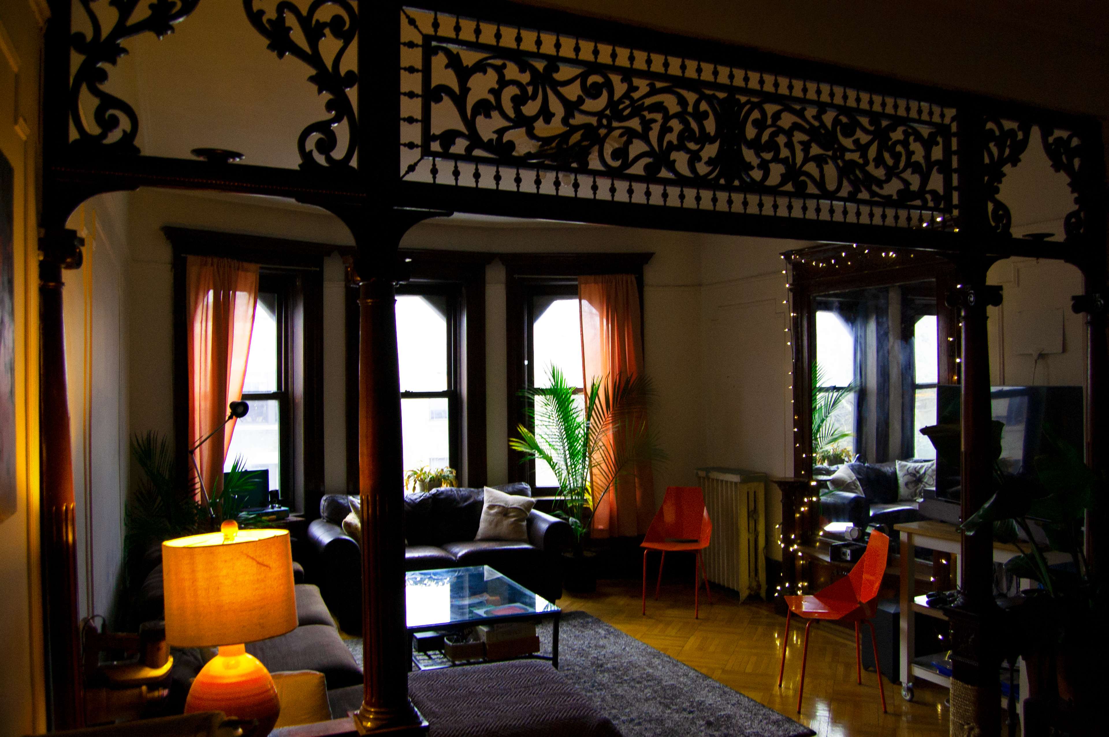 A living room with large windows, a black sofa, an orange chair, and decorative plants, featuring an ornate wooden archway.