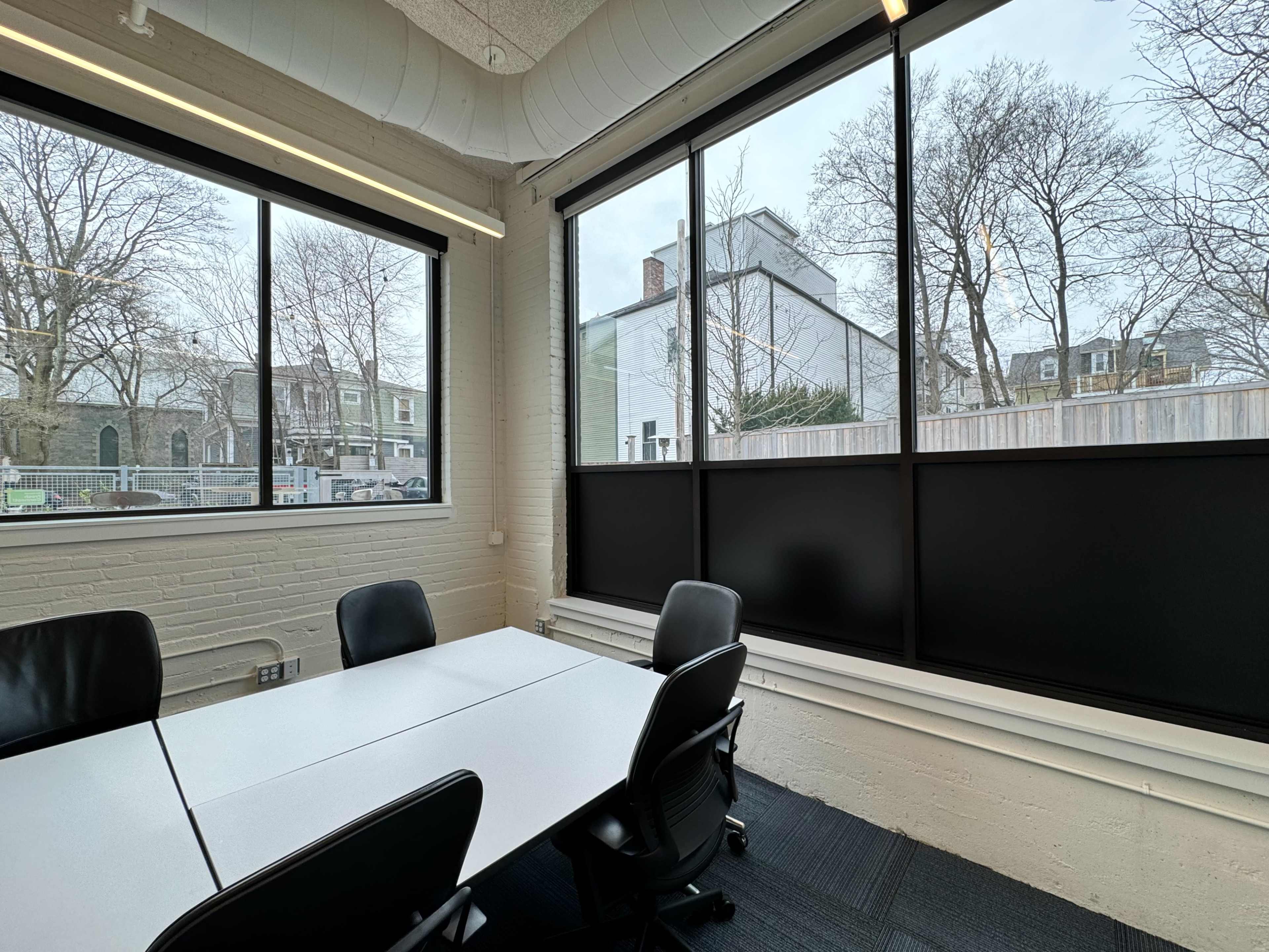 A meeting room features a white table surrounded by black chairs, with large windows revealing a view of an outdoor area and neighboring buildings.