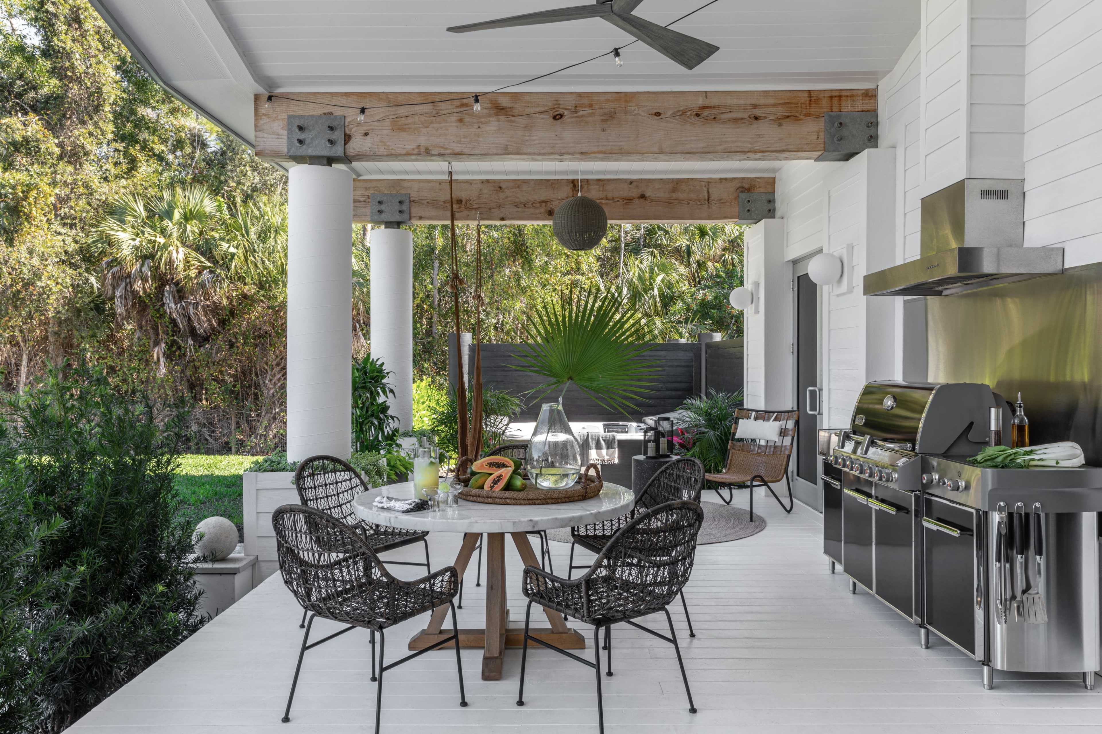 The image shows a modern outdoor kitchen and dining area featuring a circular table with chairs, a grill, and various plants in a lush backyard setting.