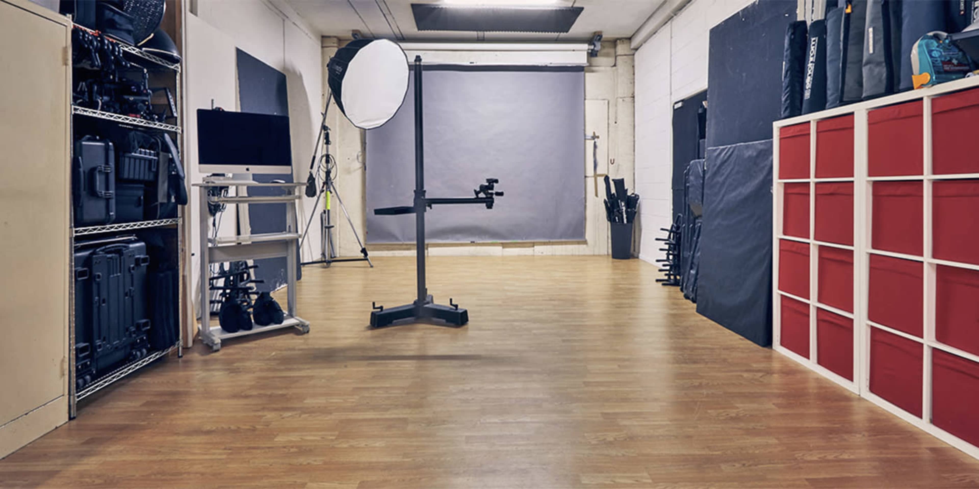 A spacious photography studio with wooden flooring, various photography equipment on shelves, and a large reflector positioned in the center.