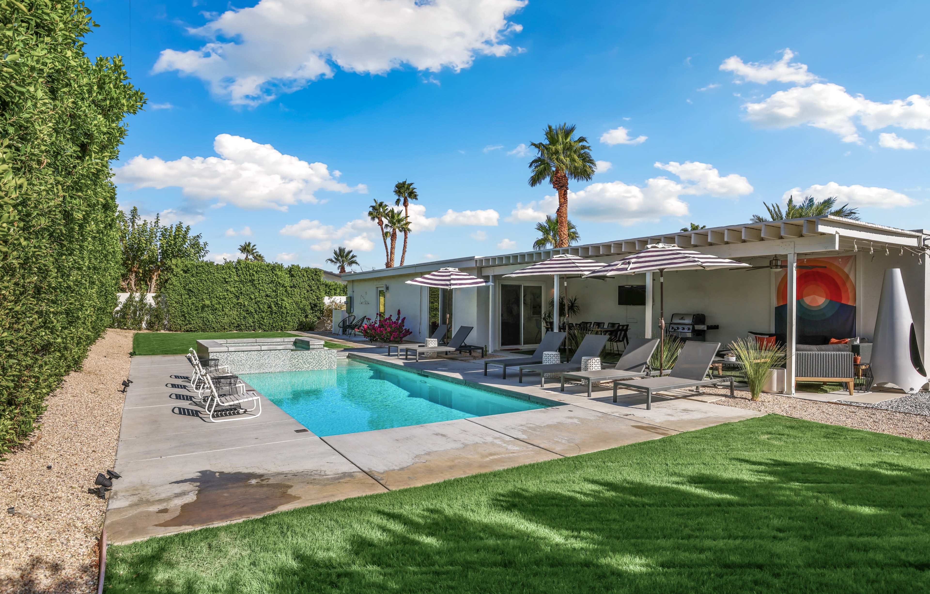 A modern backyard features a swimming pool surrounded by lounge chairs and shaded by striped umbrellas, with palm trees and green landscaping in the background.