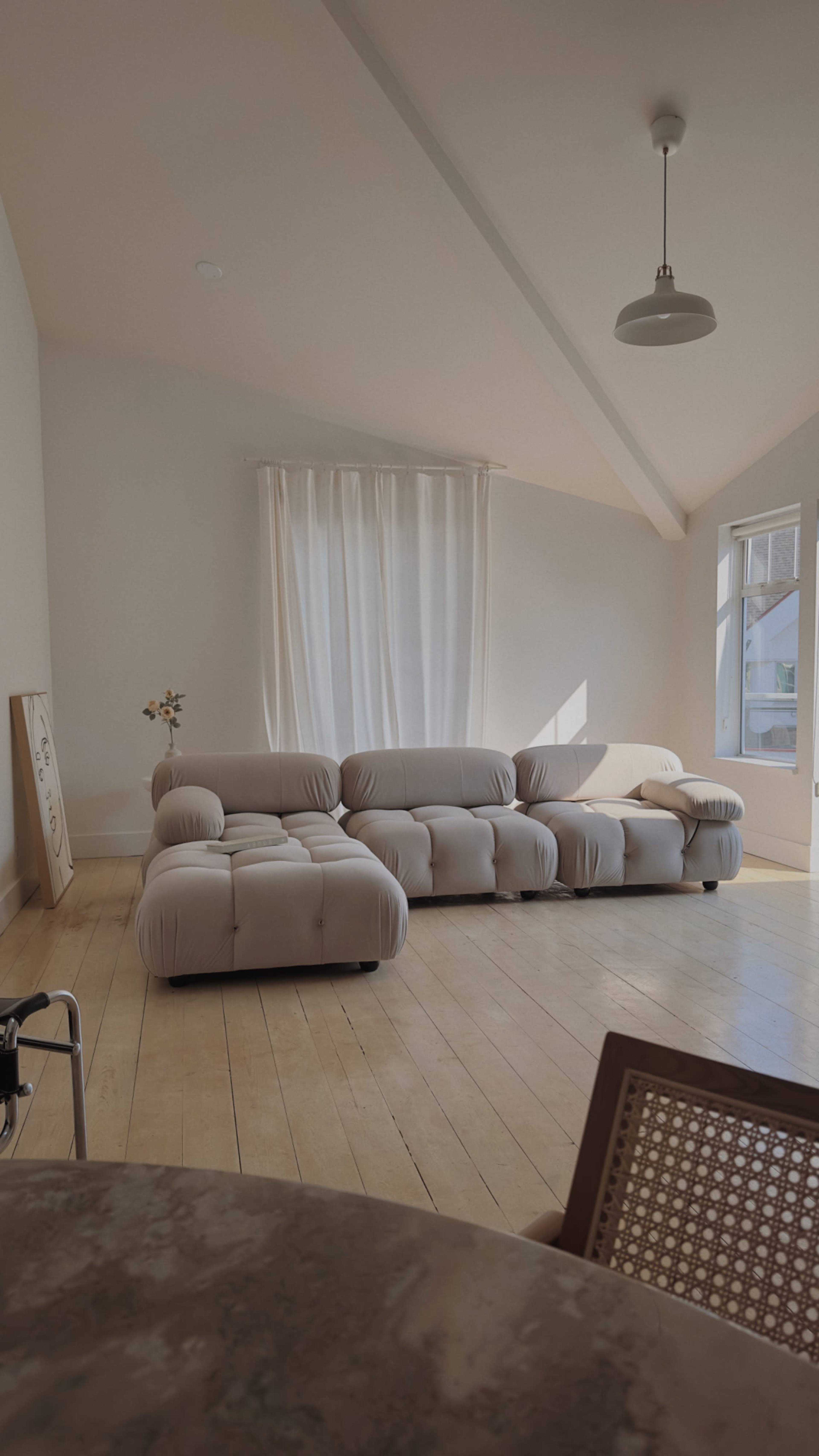 A light-colored living room with a curved beige sofa, a large window with sheer curtains, and wooden flooring.