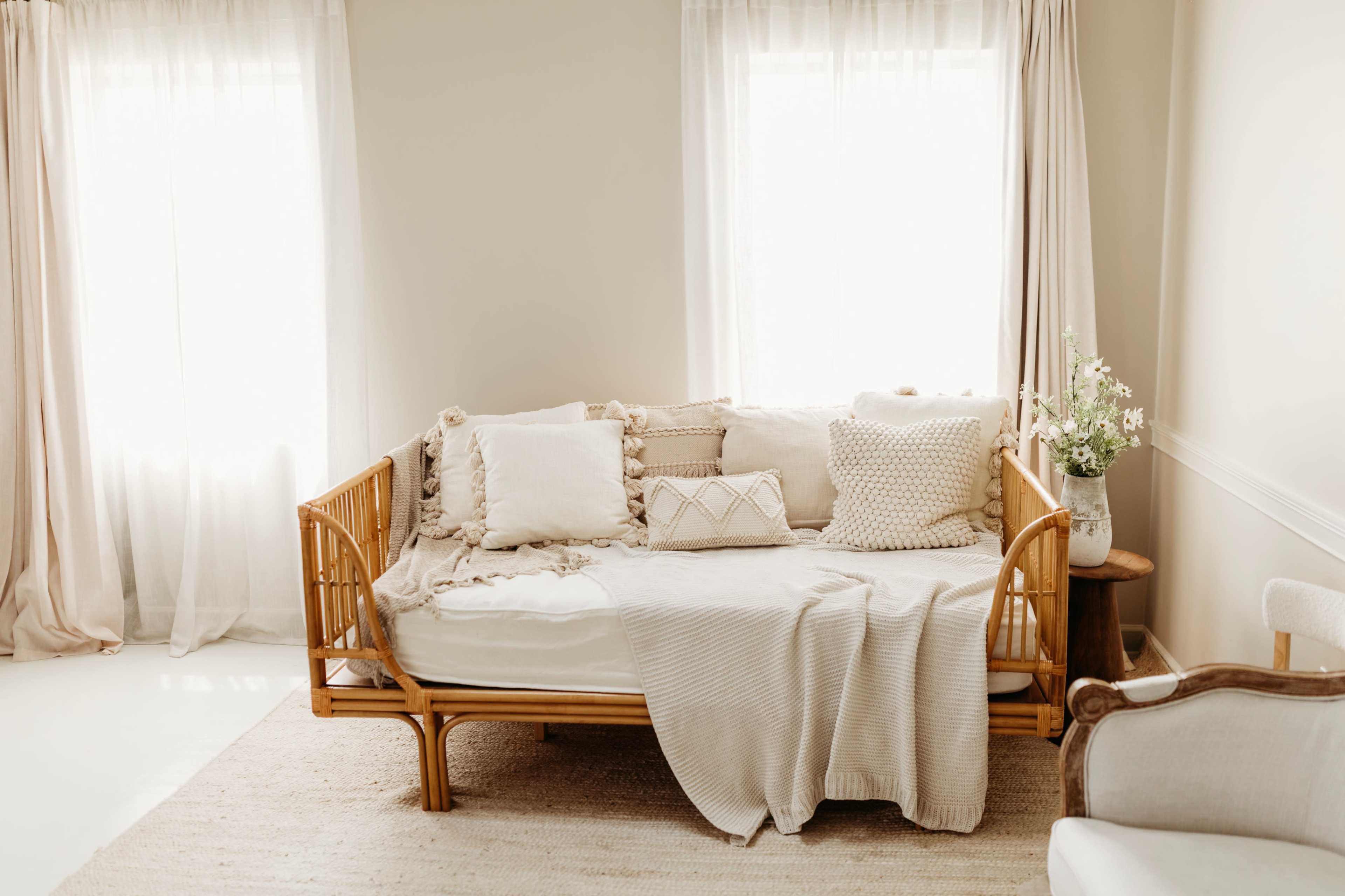 A light-filled room features a rattan daybed adorned with various textured pillows, a soft throw blanket, and sheer curtains that frame the windows.